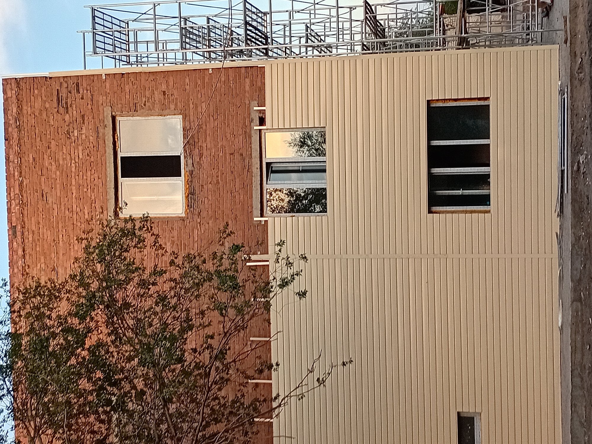The image depicts the exterior of a brick building undergoing construction or renovation. A portion of a mature tree with green foliage overhangs the upper left side of the structure. The building features a red brick upper section and a cream-colored vinyl siding lower section. There are three visible windows: one on the upper brick level with a partially open sliding pane, another on the lower siding level reflecting trees and sky, and a third on the lower siding level with multiple panes that appear dark. Scaffolding is erected on the right side of the building. The ground in front of the building is rough, with some debris visible. The sky is a pale blue, suggesting daytime. This scene is located in the city of Semey, Kazakhstan.