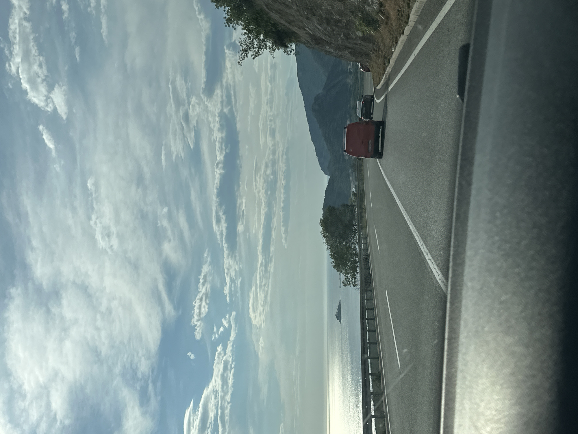 The image depicts a view from inside a car traveling on a coastal road in Kanjoši, Montenegro. The road runs parallel to the Adriatic Sea, reflecting sunlight. To the right, a guardrail separates the road from a steep slope. Two vehicles, a red van and a black car, are ahead in the same lane. The sky is predominantly blue with scattered white clouds. In the distance, a mountain range is visible. Part of the car's interior is visible on the right side of the image.