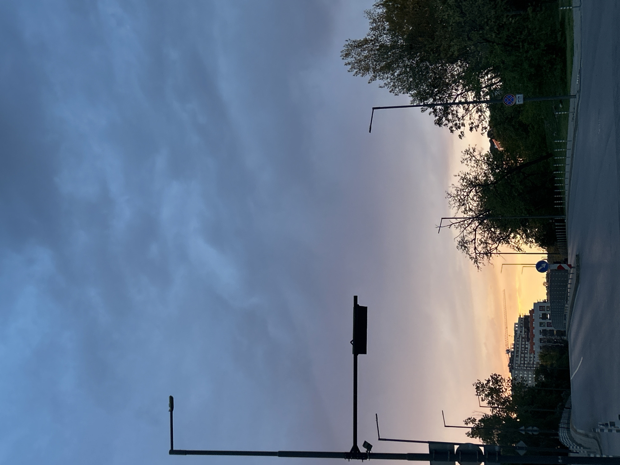 The image depicts a street scene in Sofia, Bulgaria, at dusk. The sky is predominantly gray with streaks of orange near the horizon. A road runs along the right edge of the frame, flanked by trees and buildings. Streetlights are visible along the road, casting silhouettes against the colorful sky. In the distance, a construction crane is visible amongst residential buildings. There are no people visible in the scene. A road sign is seen to indicate the direction of travel.