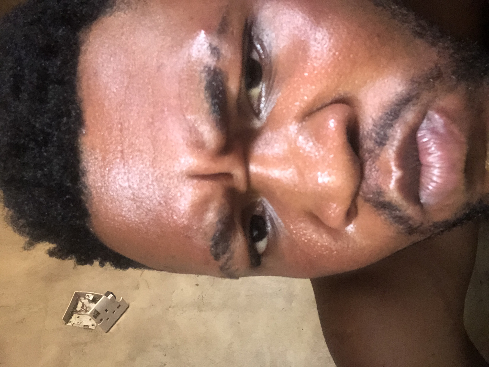 A close-up, angled self-portrait of a Black man with dark, curly hair and facial hair. His skin is shiny, likely due to sweat or oil. The background appears to be a neutral-toned floor, upon which sits an open electrical outlet box. The city and context are unknown.