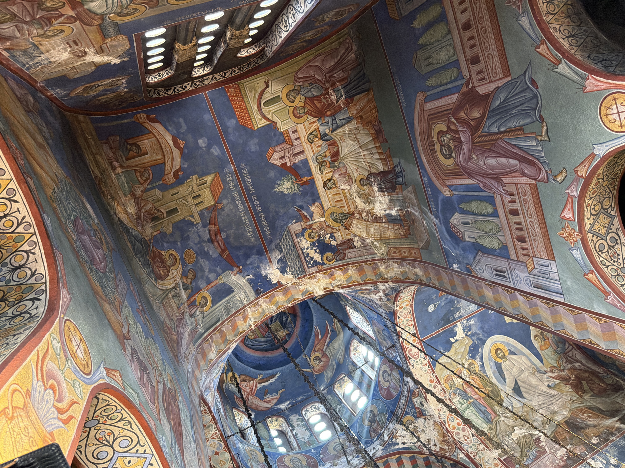 The image shows the interior of the ceiling of a church, likely in Trebinje, Bosnia and Herzegovina, decorated with intricate Byzantine-style frescoes. The frescoes depict religious scenes with figures in traditional robes, rendered in vibrant blues, reds, and golds. The ceiling curves upwards, creating a dome-like effect, and is adorned with geometric patterns and decorative borders. Chains hang down from the ceiling, possibly for chandeliers or other lighting fixtures. Sunlight streams in from arched windows with ornate grills, illuminating parts of the frescoes. The overall impression is one of rich historical and religious artistry, conveying a sense of sacredness and grandeur.