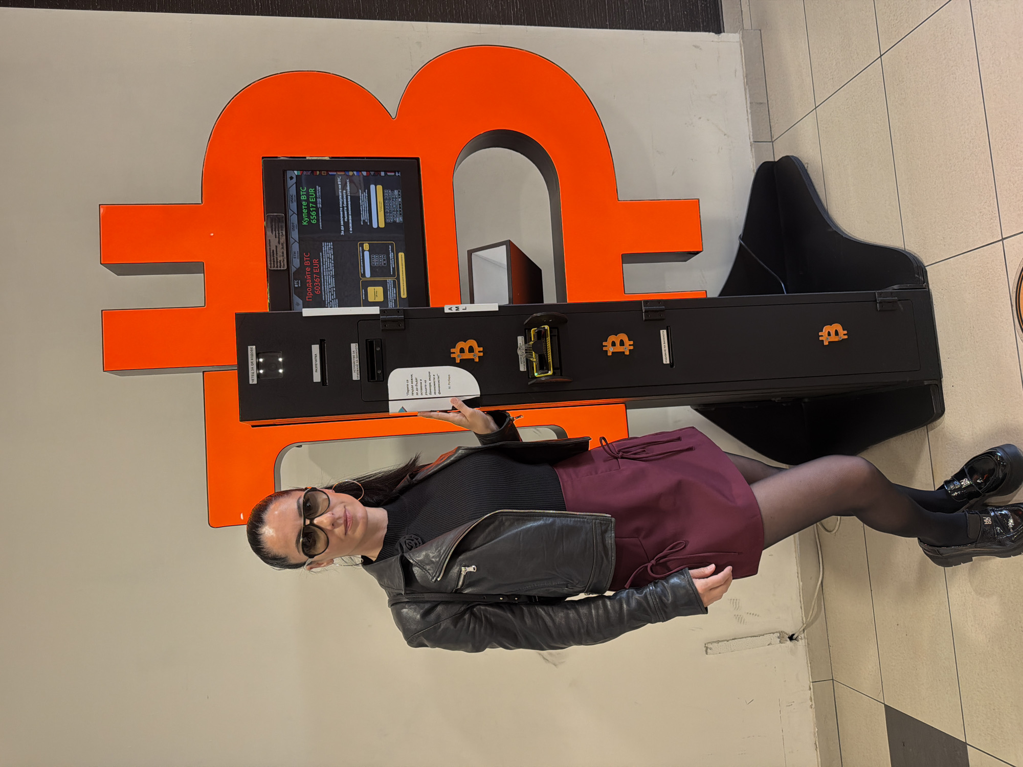 A woman stands posing next to a prominent Bitcoin ATM indoors, likely in a commercial area such as a mall in Pleven, Bulgaria. She wears a black leather jacket over a black ribbed top, a burgundy mini-skirt with ties, sheer black tights, and black patent Mary Jane-style shoes. Her dark hair is pulled back, and she wears large sunglasses and hoop earrings.
The main subject is a black cryptocurrency ATM with a screen, set against a large, orange, three-dimensional Bitcoin symbol on a light grey wall. To the right of the ATM is a black, boot-shaped decorative element. The ATM's screen displays "Продайте BTC 60367 EUR" (Sell BTC 60367 EUR) and "Купете BTC 65617 EUR" (Buy BTC 65617 EUR), indicating Bitcoin prices in Euros. Other visible text on the machine includes "ЧЕТЕЦ ЗА QR КОДОВЕ" (QR Code Reader), "ПЕЧАТКА" (Print), "AML", and "BBI". Small orange Bitcoin logos are scattered across the ATM's surface. The woman holds a piece of paper that appears to have just been dispensed from the machine, suggesting she has either completed or is demonstrating a transaction. The floor is tiled, and the area is brightly lit by artificial indoor lighting.