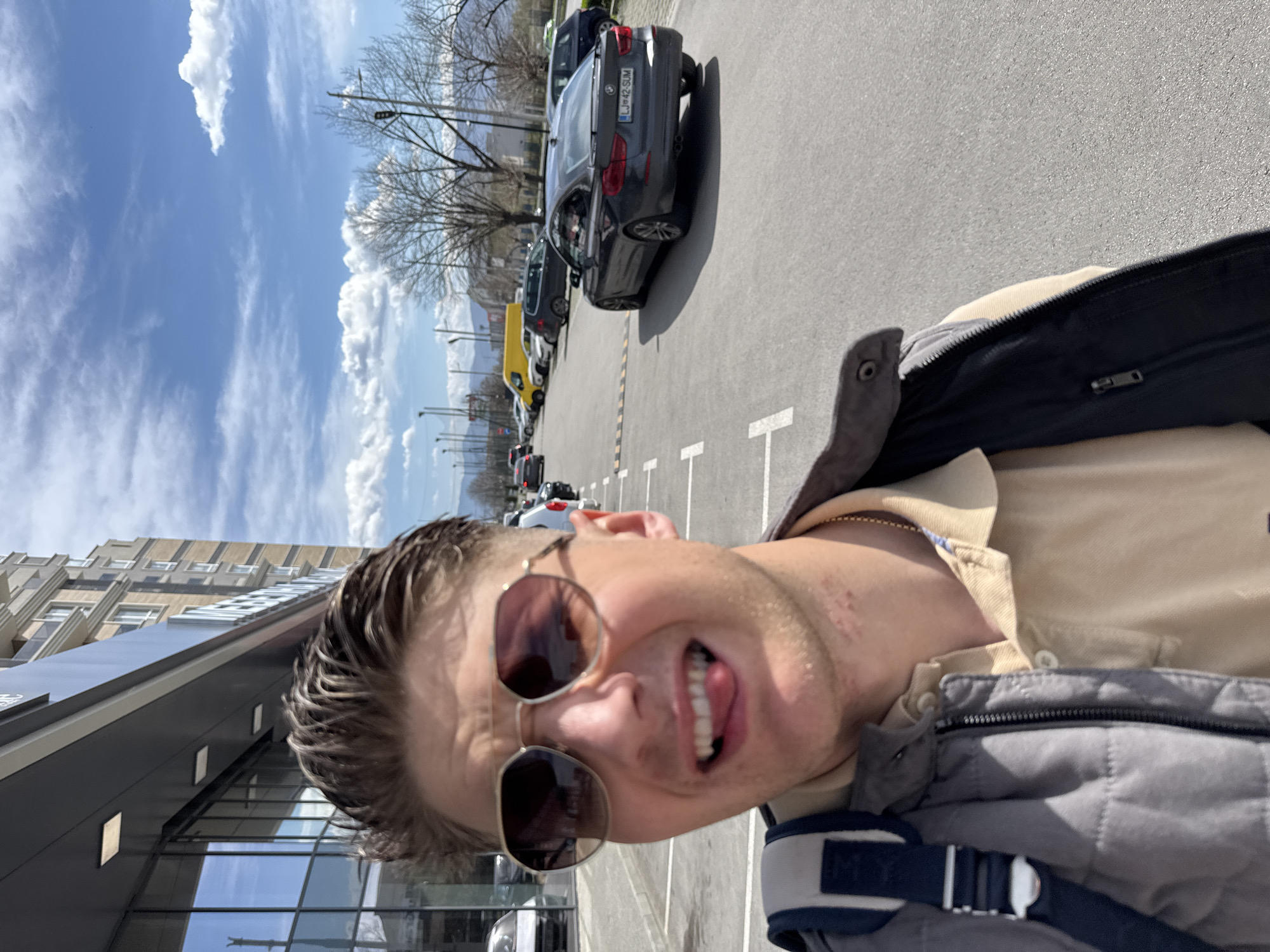 The image depicts a man wearing sunglasses and a casual outfit, including a collared shirt and vest, standing on a parking lot, sticking his tongue out at the camera. Behind him is a parking area filled with cars parked in designated spaces. The background features a building and a sky with clouds. Based on the license plate "LJ-42-SUM" visible on one of the cars, and after referencing the available context, this image was likely taken in Plovdiv, Bulgaria.