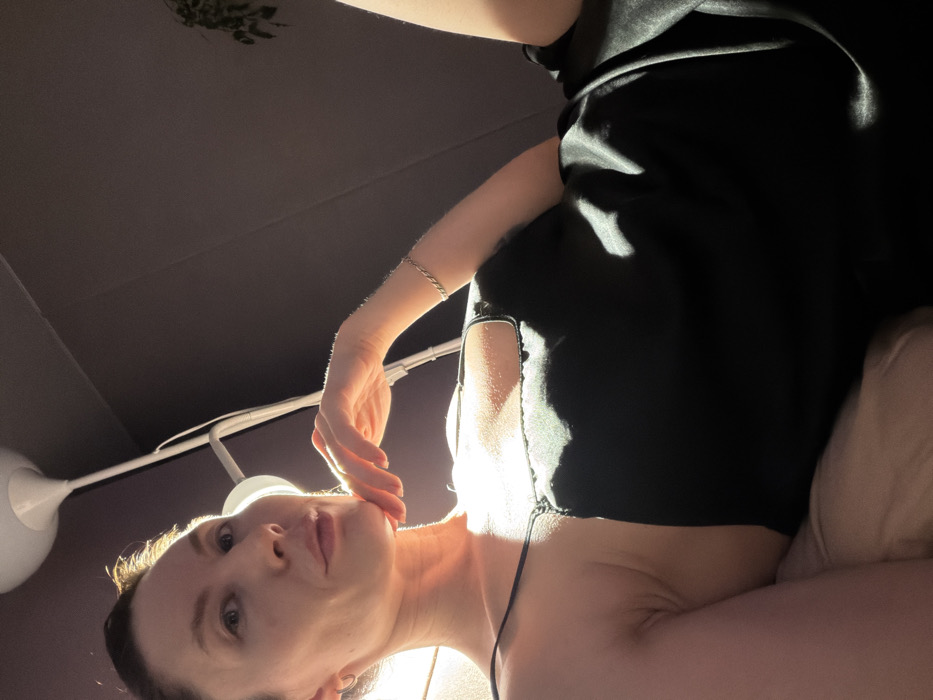 The image captures a close-up, low-angle perspective of a woman indoors, illuminated by strong, warm sunlight. The woman is positioned with her head slightly tilted, looking directly towards the camera. Her expression is calm and serene, with her lips softly parted. Her left hand is gently placed against her chin and jawline, a common pose often used for portraits. A light-colored, possibly silver, twisted bracelet is visible on her left wrist.
She is wearing a dark, likely black, top or dress with thin straps. The fabric appears smooth, possibly satin or silk, as indicated by the way the sunlight creates lustrous highlights on it. The bright, natural light strongly illuminates her face, neck, and upper chest from the left, casting dramatic highlights and soft shadows that define her features and create a glow around her. This lighting suggests it is daytime, with the sun streaming into the room.
The background is primarily a uniform dark color, resembling an indoor wall or ceiling. On the left side of the frame, a white, round-shaped lamp fixture is visible, attached to what appears to be a mounting arm or support. In the upper left corner of the dark background, a subtle, dark, and slightly blurry shape resembling foliage or a plant can be discerned. On the far right, a light-colored, soft object, possibly a pillow or cushion, is partially visible, suggesting the woman might be reclining. The overall impression is one of quiet contemplation, with the powerful natural light serving as a key artistic element. There is no visible text in the image.