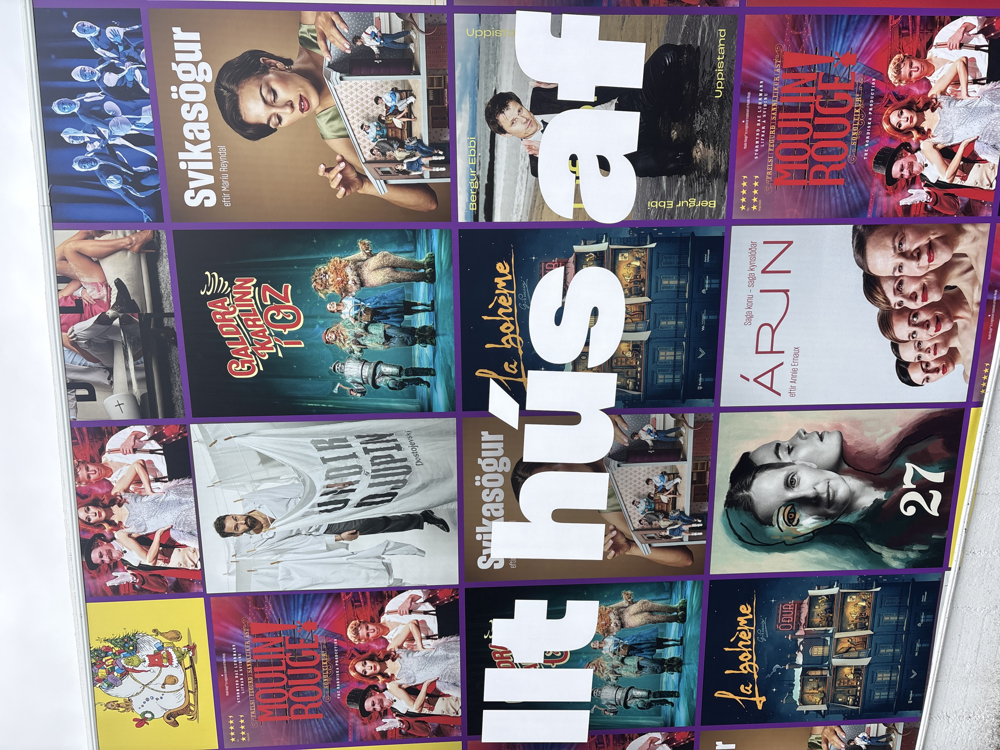 This is a photograph of a poster display for various stage productions, likely located in Reykjavik, Iceland, given the Icelandic text present. The display is arranged in a grid format, with multiple posters visible in a close-up view.
The posters feature a variety of theatrical imagery. Some depict scenes with actors in costume, such as a group in sparkly attire and another group in a stage setting with a character dressed as the Cowardly Lion from "The Wizard of Oz." Another poster shows a man hanging laundry, with text that appears to be a play title. Several posters include bold text in Icelandic, announcing titles like "Svikasögur" and "Galdra Karlinn í Óz."
One poster prominently features a close-up of a woman's face with a stylized, split artistic effect and the number "27" in large font. Another poster shows a man in a suit emerging from water. Several posters are for a production titled "Moulin Rouge," featuring the show's iconic red lettering and cast members. The overall impression is a vibrant and diverse collection of promotional materials for the performing arts.