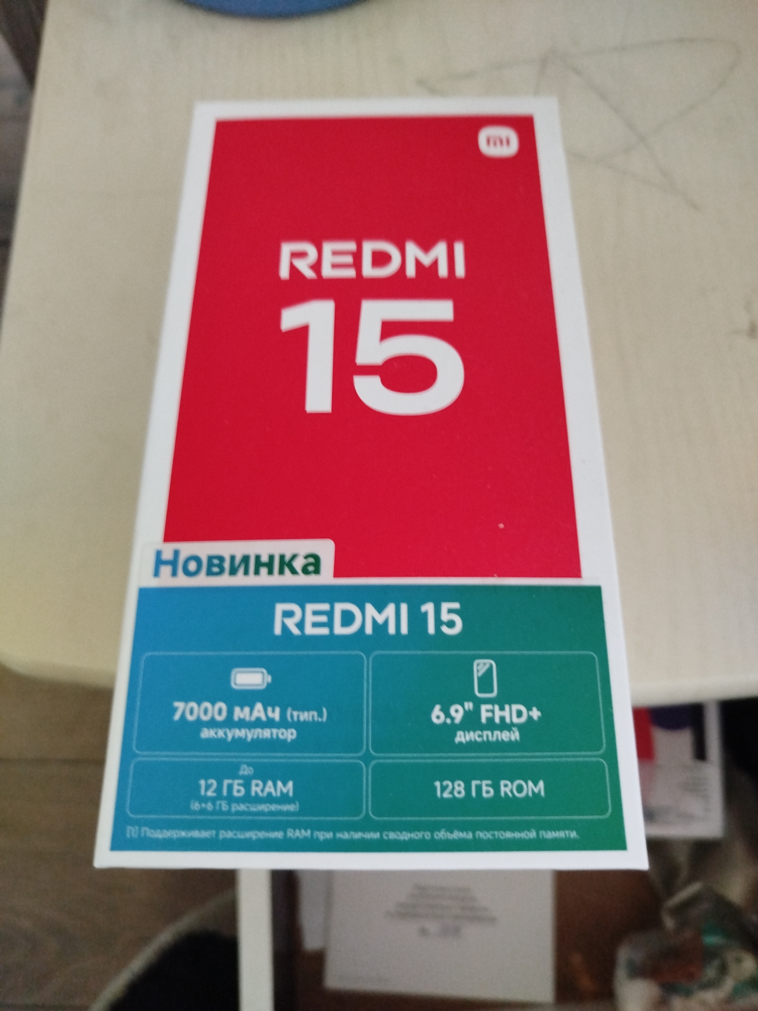 A red and white box for a Redmi 15 smartphone is displayed. The box prominently features the brand name "REDMI" and the number "15" in large white font on a red background. Below this, the word "Новинка" (New Arrival) is written in blue. The bottom section of the box is green and lists specifications of the phone, including "7000 мАч (тип.) аккумулятор" (7000 mAh (typical) battery), "12 ГБ RAM" (12 GB RAM), "6.9" FHD+ дисплей" (6.9" FHD+ display), and "128 ГБ ROM" (128 GB ROM). The box appears to be sitting on a surface that is partially visible at the top and bottom of the image. The overall impression is that of product packaging, likely for retail display or sale. There is no indication of the location being in Крывасельскі сельскі Савет, Belarus.