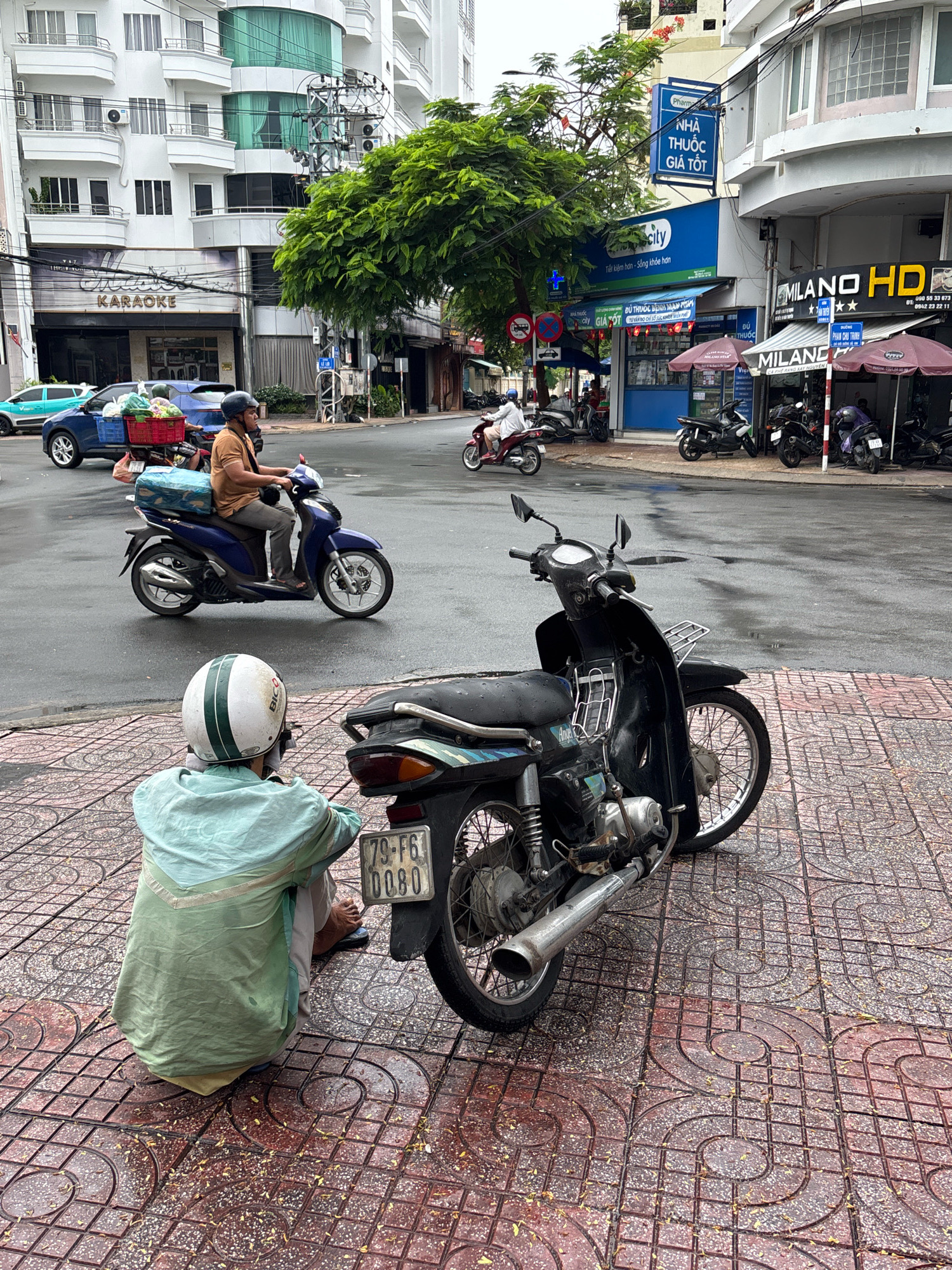 In Nha Trang, Vietnam, a street scene unfolds with a person squatting on a red tile sidewalk next to a black motorcycle. The person, wearing a light green shirt and a white and green helmet, is facing away from the camera. The motorcycle, a black scooter with a license plate reading "79-F6 0080," is parked on the sidewalk.
In the background, a blue car with a red basket full of produce is on the street, and a person in a brown shirt is riding a purple scooter, also carrying a large blue package. Several other motorcycles are parked in front of shops. The buildings in the background include "KARAOKE" signage and other businesses. The street is wet, suggesting recent rain. The overall scene depicts a typical urban street environment in Vietnam.