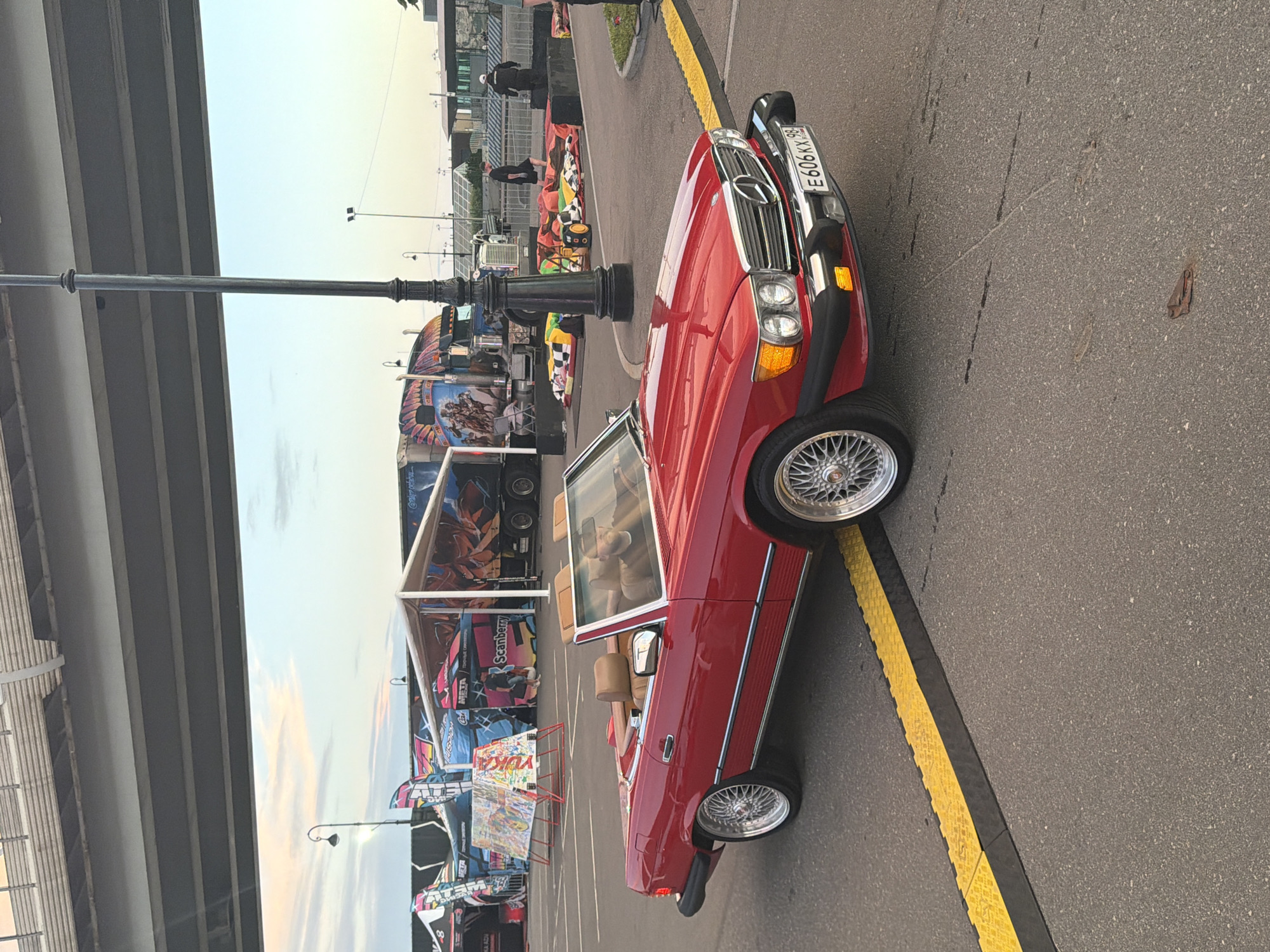 A red Mercedes-Benz SL convertible with a tan interior is parked on asphalt next to a yellow and black speed bump. The car has chrome wheels and a license plate that reads "E 606 KX 98". In the background, there are tents, a semi-truck with artwork, and a lamppost. The sky is a light blue with some pink and orange hues from the sunset. A man wearing a dark shirt and a cap is sitting in the driver's seat. The scene appears to be an outdoor event or fairgrounds.