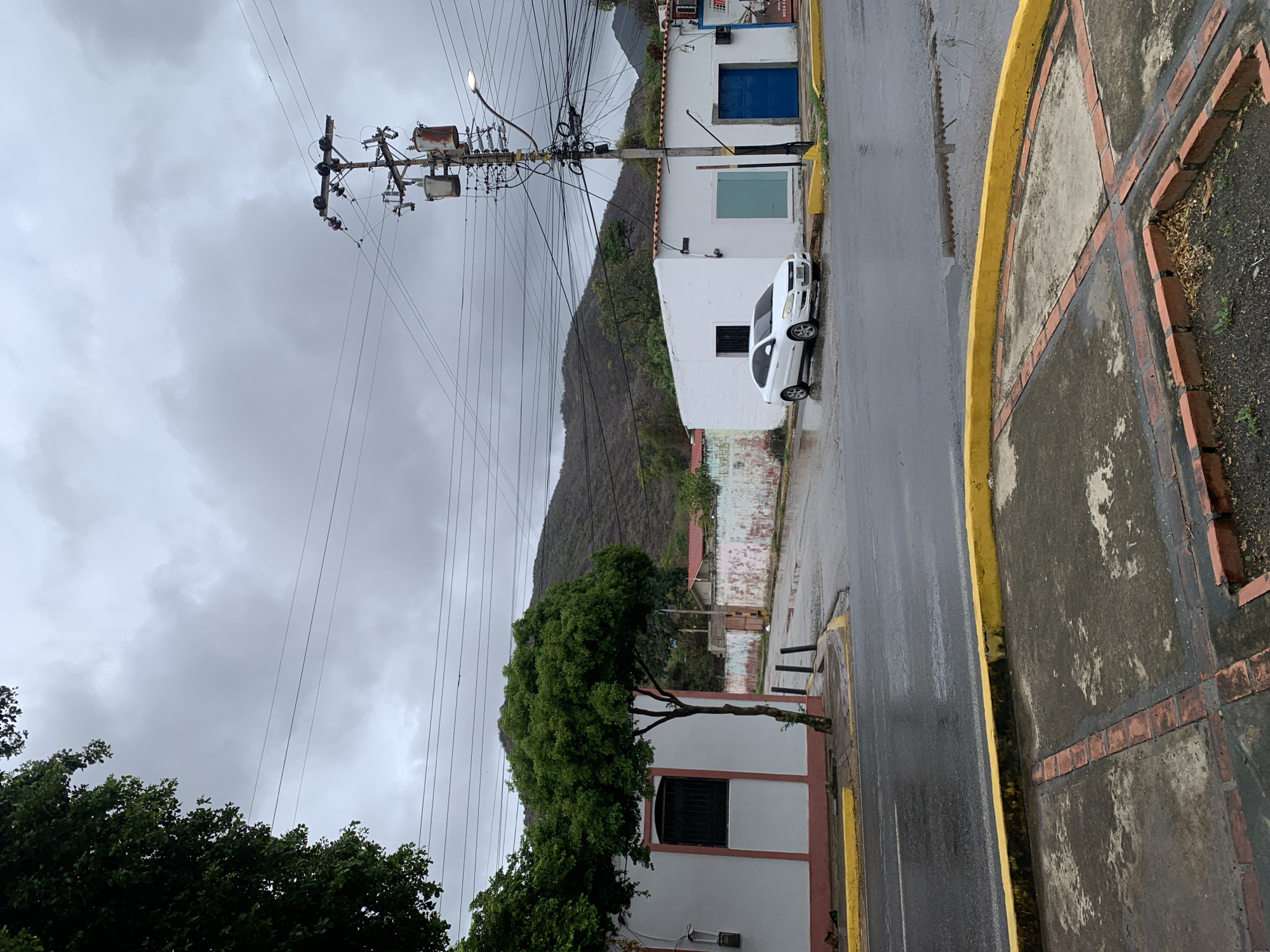 The image depicts a street scene in Santa Ana, Venezuela, under an overcast sky. A white four-door sedan is parked on the wet asphalt in front of a white building with a blue door. Adjacent to it is another white building with a teal-colored window. A mature tree with dense green foliage stands near a white building with a red trim and a barred window. In the background, a brown, tree-covered hill rises. Utility poles with numerous wires and transformers are visible. The road and sidewalk are damp, with puddles reflecting the cloudy sky. A yellow-painted curb delineates the sidewalk from the street.