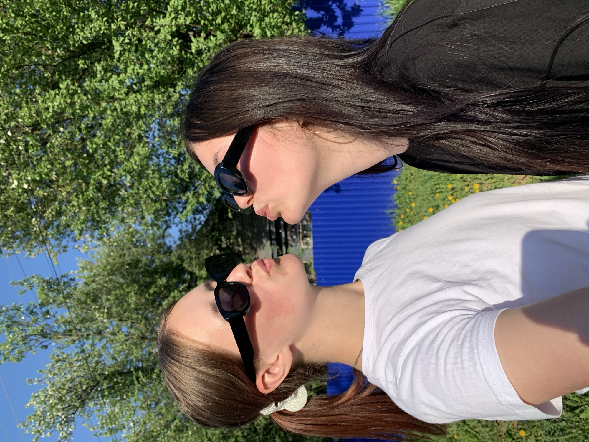 Two women, wearing sunglasses, are positioned close together outdoors. The woman on the right wears a black shirt and has long, dark brown hair. The woman on the left wears a white shirt and has light brown hair pulled back in a ponytail secured with a white hair accessory. The background includes trees and what appears to be a blue fence. The image seems to be taken on a sunny day. There is no way to confirm the location of Фоки, Russia.