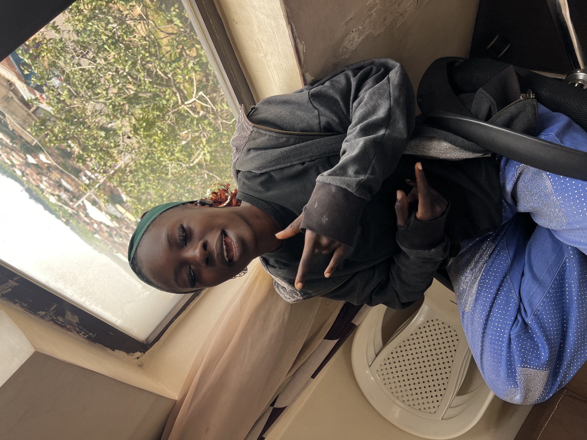 A Black woman wearing a green headscarf, a grey zip-up hoodie over a black t-shirt, and blue jeans adorned with silver rhinestones, sits in a chair. She is looking towards the camera with her mouth slightly open and her right hand held up, fingers pointed in a gesture. She is positioned indoors, near a window that offers a view of a cityscape with buildings and trees, likely in Jos, Nigeria. A white plastic chair and a portion of a desk chair are visible in the foreground. The lighting suggests it is daytime, and the environment appears somewhat rustic.