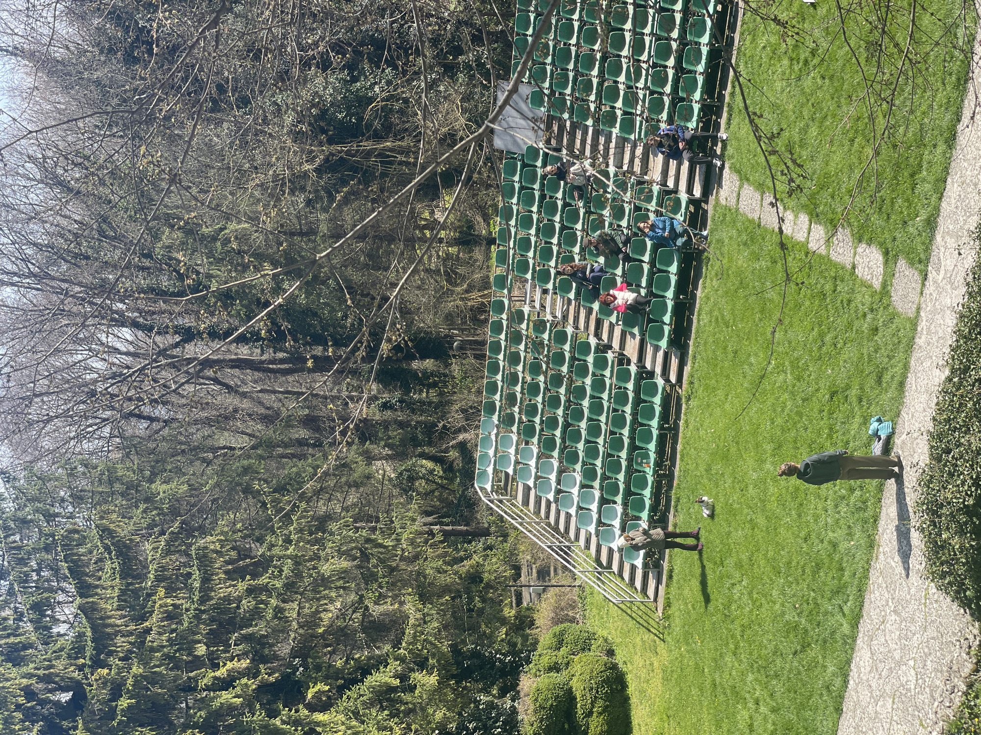 The image shows an outdoor scene featuring stadium-style green seating placed on a grassy area. Several people are seated in the stands, and two individuals are standing on the ground level near the seating. Adjacent to the seating area is a paved path with overgrown stone patterns, with trees and bushes surrounding the area. Based on visual features and the location provided, the scene might depict a part of the Sea Garden in Varna, Bulgaria.