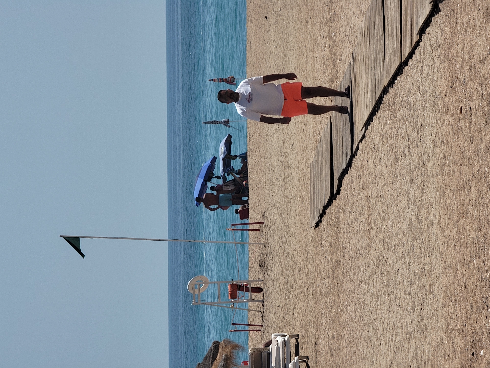 A full-frame, eye-level, outdoor medium shot captures a sandy beach in Quarteira, Portugal, on a bright, sunny day with a clear blue sky. In the foreground, a man walks on a wooden boardwalk. He is middle-aged, with dark hair and a mustache, wearing a white t-shirt with a graphic print and orange shorts. He is walking towards the camera with his arms at his sides, his face neutral. The boardwalk extends into the background, curving slightly to the left.
The middle ground features the sandy beach with a few beach chairs and umbrellas. A lifeguard stand with a red life preserver is visible. In the background, the ocean is a vibrant blue, with gentle waves. A few people are visible in the distance, some under umbrellas. A dark green flag flies from a tall pole on the left side of the image, indicating good swimming conditions. The overall impression is a peaceful beach scene on a warm day.