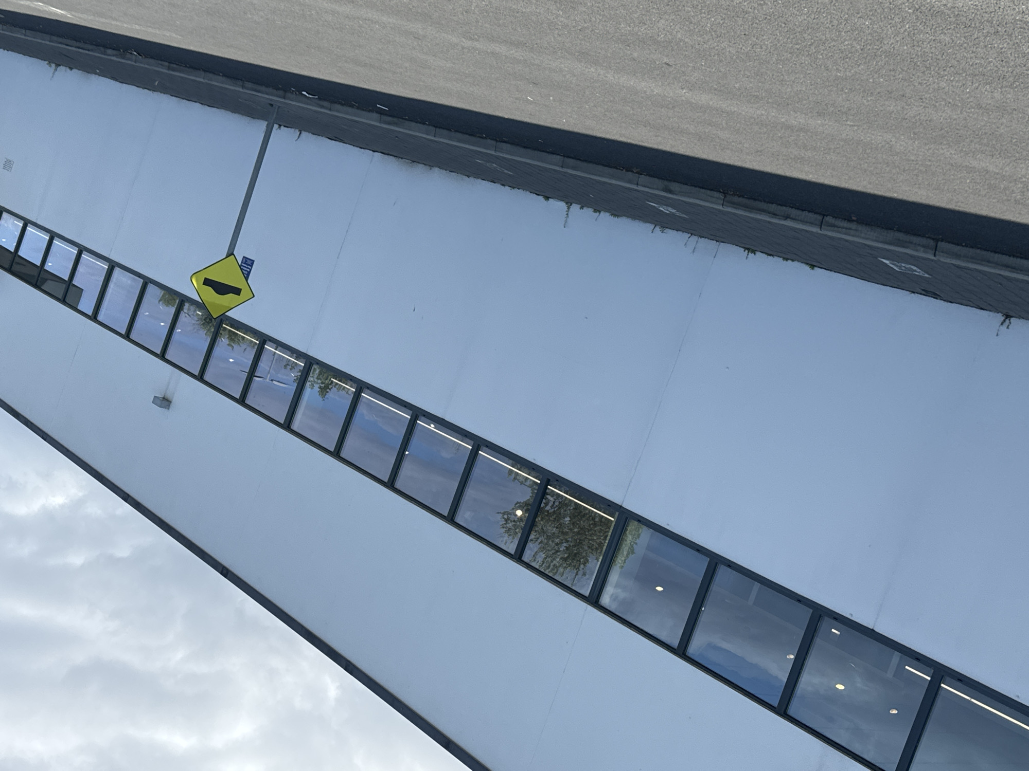 The image shows a long, white, modern building with a continuous band of windows running diagonally across its facade. The windows reflect a cloudy sky and some trees. In front of the building, there is a paved area with a sidewalk, and beyond that, a road. A yellow diamond-shaped traffic sign indicating a speed bump is visible on a pole in front of the building. The scene appears to be an industrial or commercial area on an overcast day. The image was taken in the city of Waterford, Ireland.