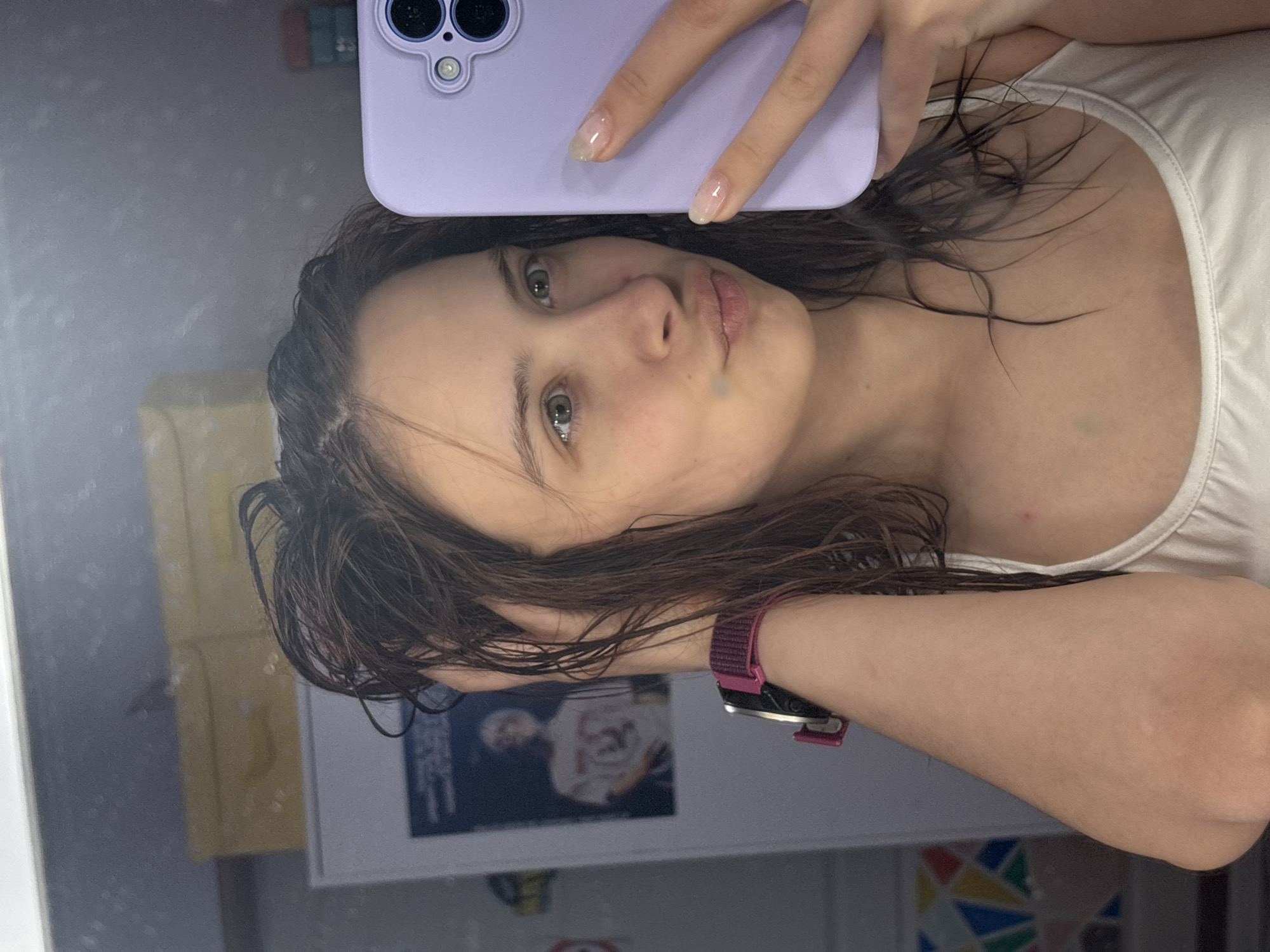 A young woman with wet, dark brown hair takes a selfie in what appears to be a bathroom. She is holding a purple phone up to her face and wearing a white tank top. The background includes a yellow object, a framed poster of a person wearing a white shirt with number 23 and a sign with colors triangles. Based on location metadata, the image was taken in the city of Прикамский, Russia. The woman is wearing a maroon wrist watch. Her nails are manicured with a clear coat.
