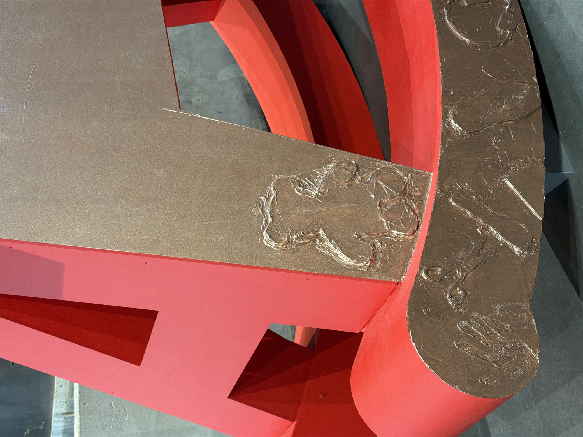This image captures a detail of a large, abstract sculpture, likely situated indoors in a gallery or museum setting. The artwork is composed of bold, geometric, and curved elements in contrasting colors and textures.
The primary colors are a vibrant coral red and a metallic bronze. Large, smooth planes and angular structures are painted in the coral red, with some sections showing darker red recesses that create a sense of depth and shadow. Intersecting with and overlying these red forms are metallic bronze elements. These bronze surfaces are highly textured, featuring raised, abstract patterns and organic-like shapes that resemble crude drawings, swirls, or impressions. On the large, curved bronze section to the right, some of these textures might suggest handprints or claw marks, adding to an raw, tactile quality.
The sculpture's composition involves an interplay of straight lines, sharp angles, and sweeping curves. A prominent flat bronze surface extends across the top and mid-left, adorned with more of the textured patterns, before meeting a vertical, curved bronze element on the right. Below these, the red structures form complex, multi-layered shapes, with cut-outs revealing deeper red interior sections.
The background is a muted, dark gray or concrete-like wall, which allows the vibrant colors and intricate textures of the sculpture to stand out. The lighting appears even, suggesting an indoor environment with controlled artificial illumination. There is no discernible text visible within the image. The overall impression is one of modern abstract art, with a focus on form, color, and tactile surface qualities. Given the context, this artwork is located in Moscow, Russia.