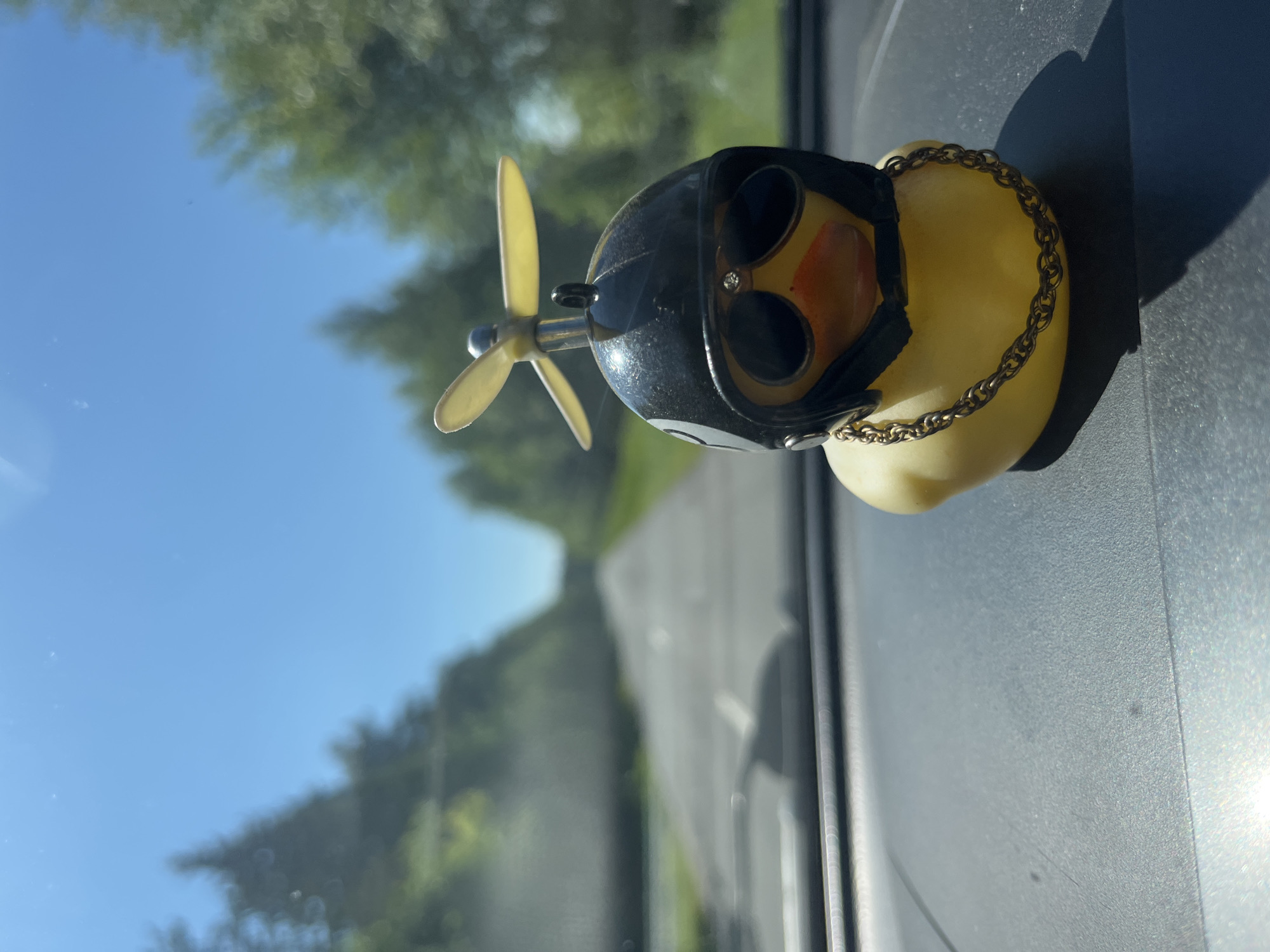 A close-up, eye-level shot shows a novelty yellow rubber duck sitting on a car's dashboard. The duck is adorned with a black helmet featuring a propeller on top, dark sunglasses with rhinestones, and a gold chain around its neck. The background is blurred, revealing a sunlit road with trees lining both sides, suggesting a scenic drive. The car's interior, particularly the textured dashboard, is in focus in the foreground. The scene is set in an outdoor, daytime environment under a clear blue sky. The location is not specified as Simekeikasy, Russia, or any other specific city or country.