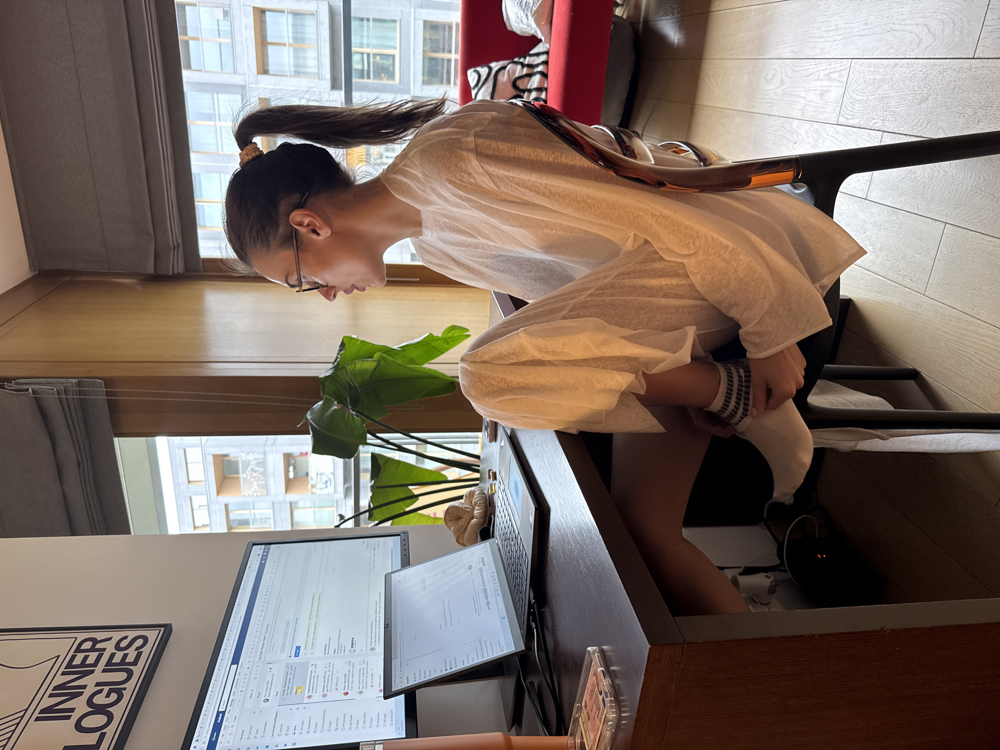 A woman with brown hair pulled into a ponytail sits at a desk in an office space, facing forward and turned to the left. She wears glasses and a white, oversized shirt and has one foot crossed over her opposite knee. A monitor screen displays an email program. A smaller screen sits next to it. The desk is wooden, and a plant sits beside the screens. An "Inner Logues" poster is on the left wall. Windows are behind her, showing an exterior cityscape. A red chair and pillows are visible to the right of her. The location is a city with tall buildings.