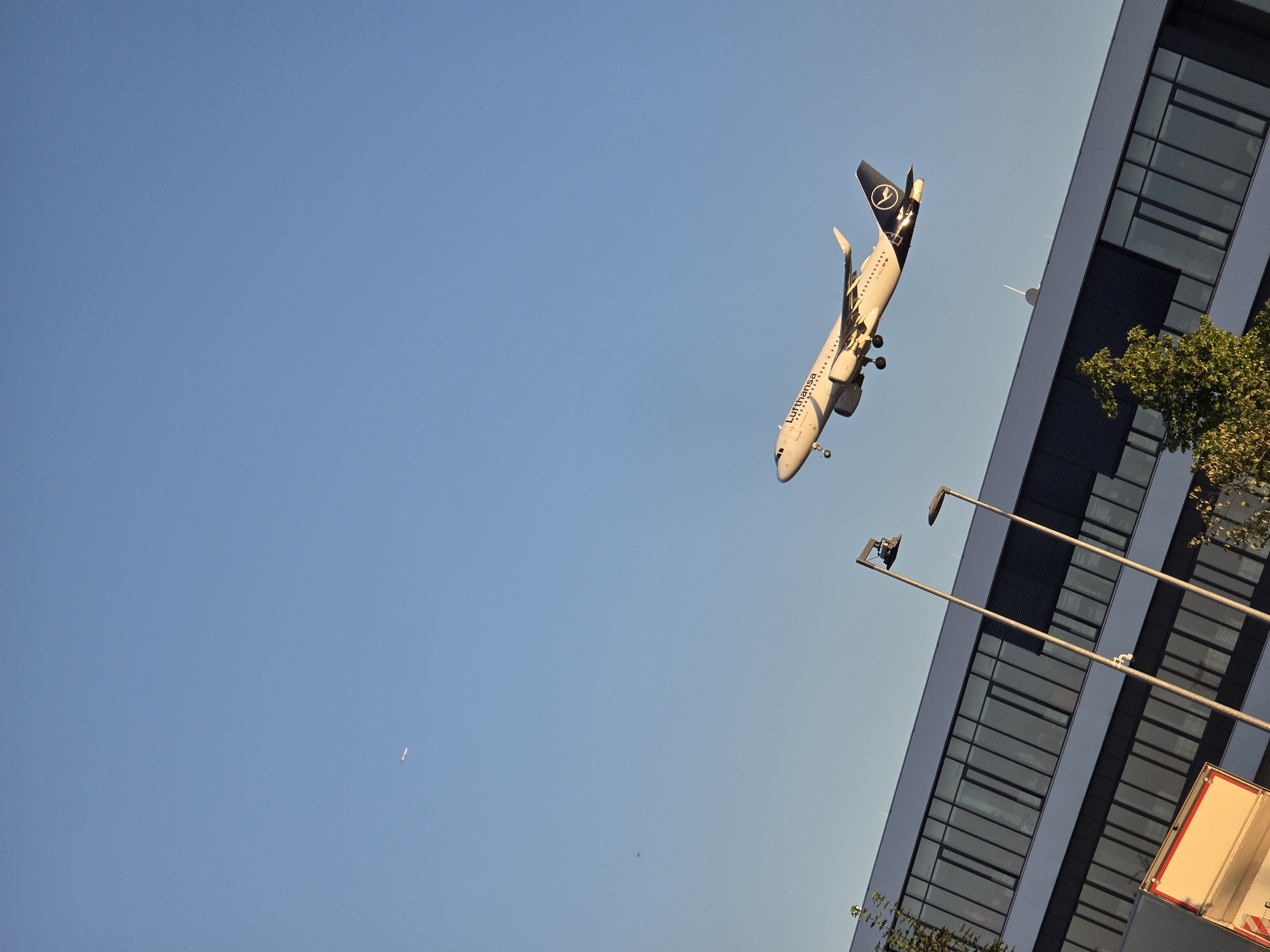 This image captures a Lufthansa airplane ascending into a clear blue sky, with a modern, angular building forming the foreground and midground. The building's facade is characterized by large, rectangular windows with dark frames, reflecting the sky. Two tall, slender lamp posts stand in front of the building, angled upwards. The airplane, a white passenger jet with the Lufthansa logo on its tail, is positioned in the upper right quadrant of the image, appearing to be in the process of taking off or climbing. In the far upper left of the sky, a smaller, more distant aircraft is visible. The overall lighting suggests it is either early morning or late afternoon, given the warm glow on the building and the long shadows cast by the lamp posts. The image is taken from a low angle, looking up towards the sky and the ascending aircraft. This scene is consistent with the airport environment of Kelsterbach, Germany, which is located adjacent to Frankfurt Airport.