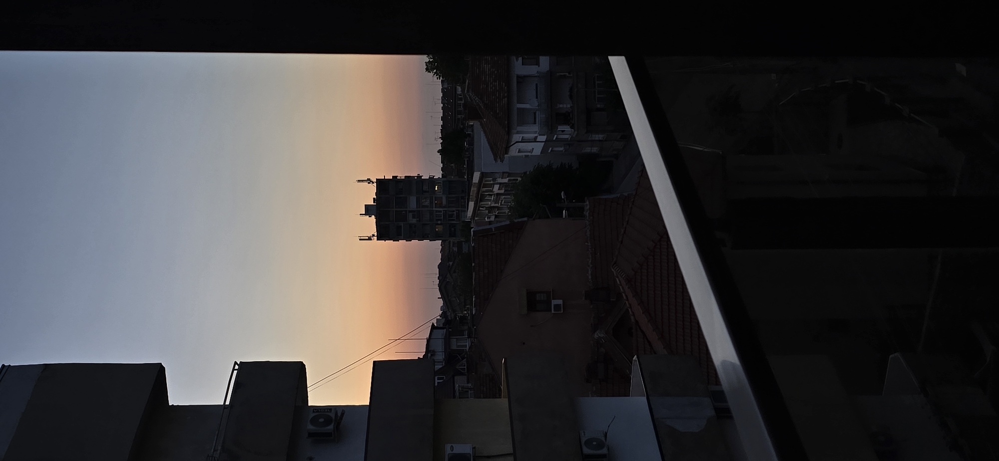 The image captures an urban landscape at dusk from an elevated perspective, likely a balcony or window, in the city of Unknown, Unknown. The sky transitions from a pale blue at the zenith to a warm orange and pink hue along the horizon, indicative of sunset.
In the immediate foreground, a dark, metallic balcony railing extends diagonally from the lower left towards the lower right. To the far left, a portion of a multi-story building, from which the photograph appears to be taken, is visible in shadow, showing multiple balconies and outdoor air conditioning units. To the far right, a dark vertical structure frames the scene.
The midground is dominated by a dense collection of buildings, primarily residential, with terracotta-tiled roofs. These structures are largely silhouetted against the diminishing daylight. A prominent feature is a taller, multi-story building centrally located, characterized by a somewhat modular or brutalist architectural style, with two distinct antenna-like structures on its roof. Power lines or communication cables are visible stretching across the mid-distance. Further into the midground, additional buildings, some with multiple stories and visible windows, line what appears to be a street or alleyway. Minimal foliage from trees is interspersed among the buildings.
The background consists of the distant urban horizon line under the vibrant sunset sky. No individuals are visible in the scene.