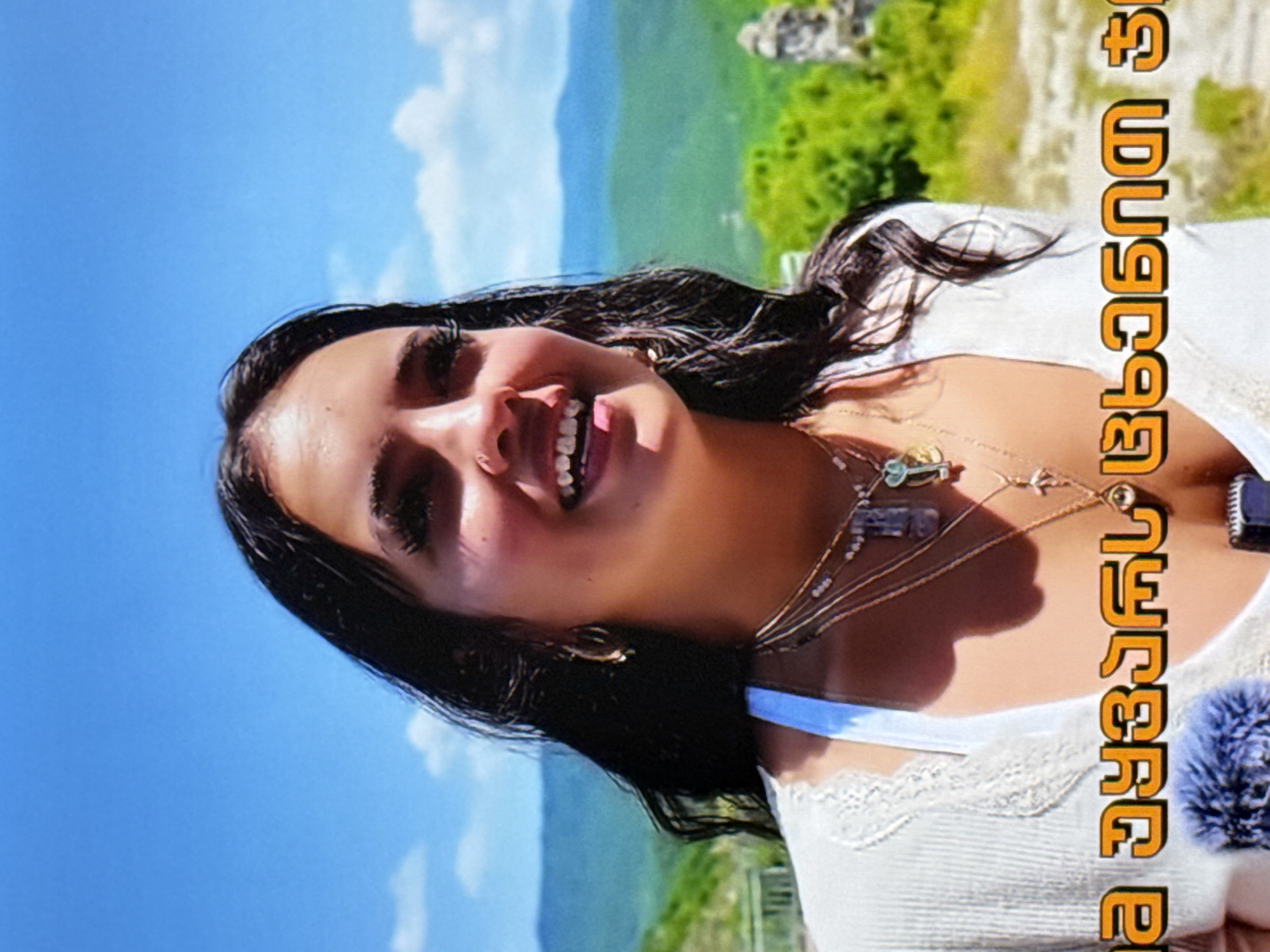 A woman with dark, wavy hair smiles and looks to her right. She is wearing a white, lace-trimmed top and multiple necklaces, including one with a key pendant and another with a faceted purple stone. She has hoop earrings and her makeup includes dramatic false eyelashes. The background is a bright blue sky with some white clouds, and a lush, green mountainous landscape. Text in an orange, stylized font appears at the bottom of the image.