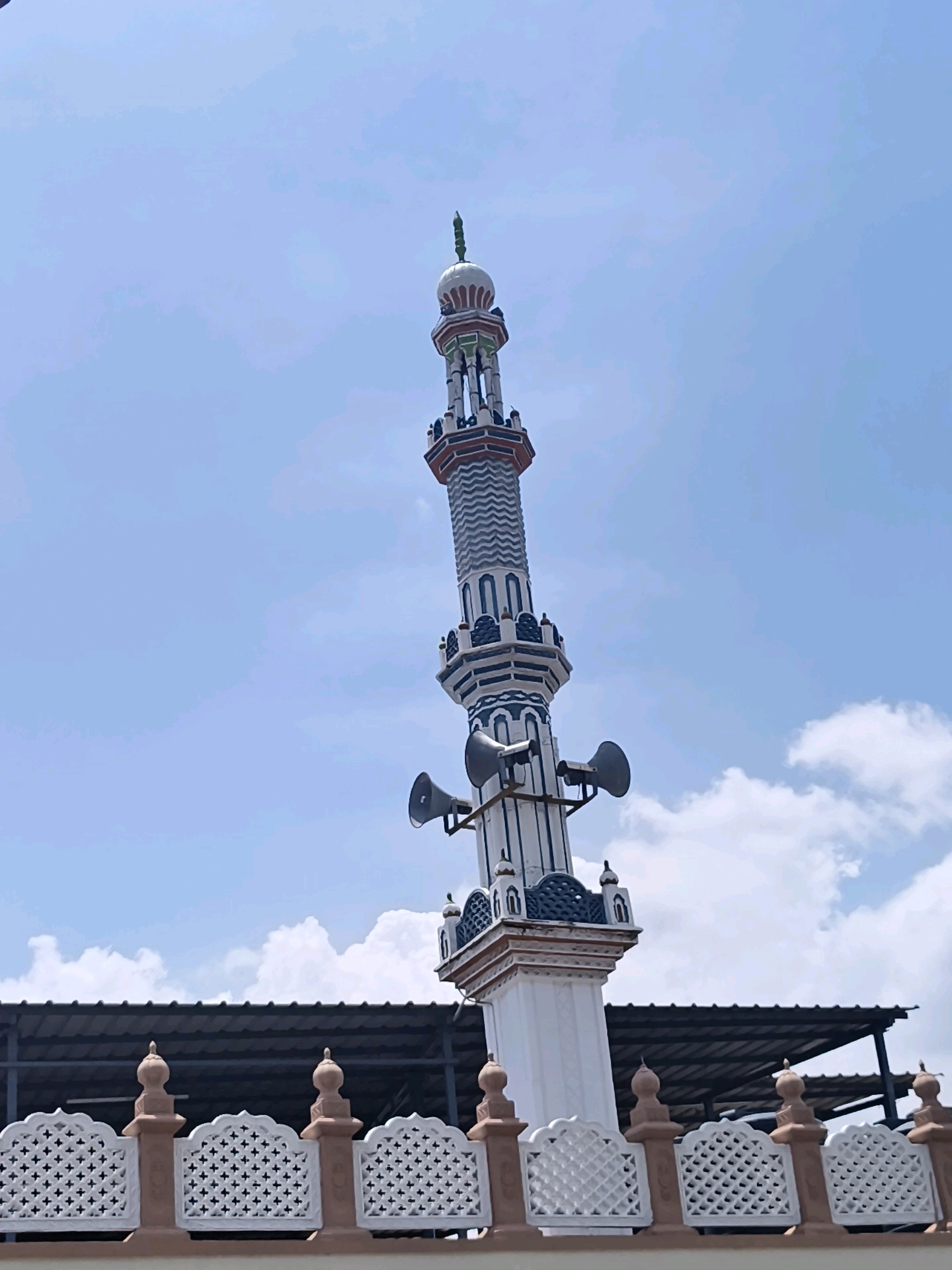 The image shows a minaret of a mosque against a blue sky with some clouds. The minaret is tall and slender, with a white base and a decorative upper section in white, blue, and red. Two loudspeakers are mounted on the minaret. In the foreground, a decorative white and brown fence runs horizontally across the frame, with a dark-colored roof structure visible behind it. The image appears to be taken outdoors during the daytime.