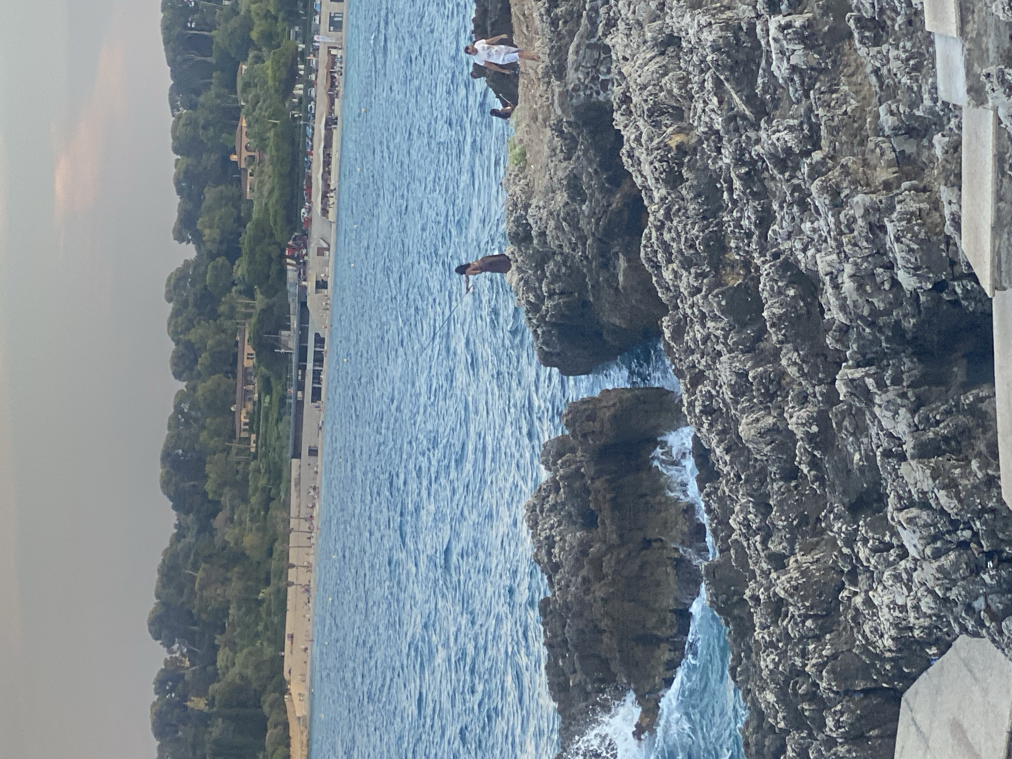 This image depicts a rocky coastline with the Mediterranean Sea in the background, likely in Antibes, France. In the foreground, rough, gray rocks form the shoreline, with waves crashing against them. Further out in the sea, three people are visible on the rocks. One individual, wearing a dark dress, stands with a fishing rod, appearing to cast a line into the water. Another person, in a white shirt and shorts, walks away from the viewer. A third, less distinct figure is seated on the rocks. In the distance, a sandy beach with scattered umbrellas and beachgoers lines the shore. Behind the beach, a lush green hillside is dotted with buildings, characteristic of the Antibes landscape. The sky is overcast, suggesting a calm, possibly late afternoon or early evening scene.