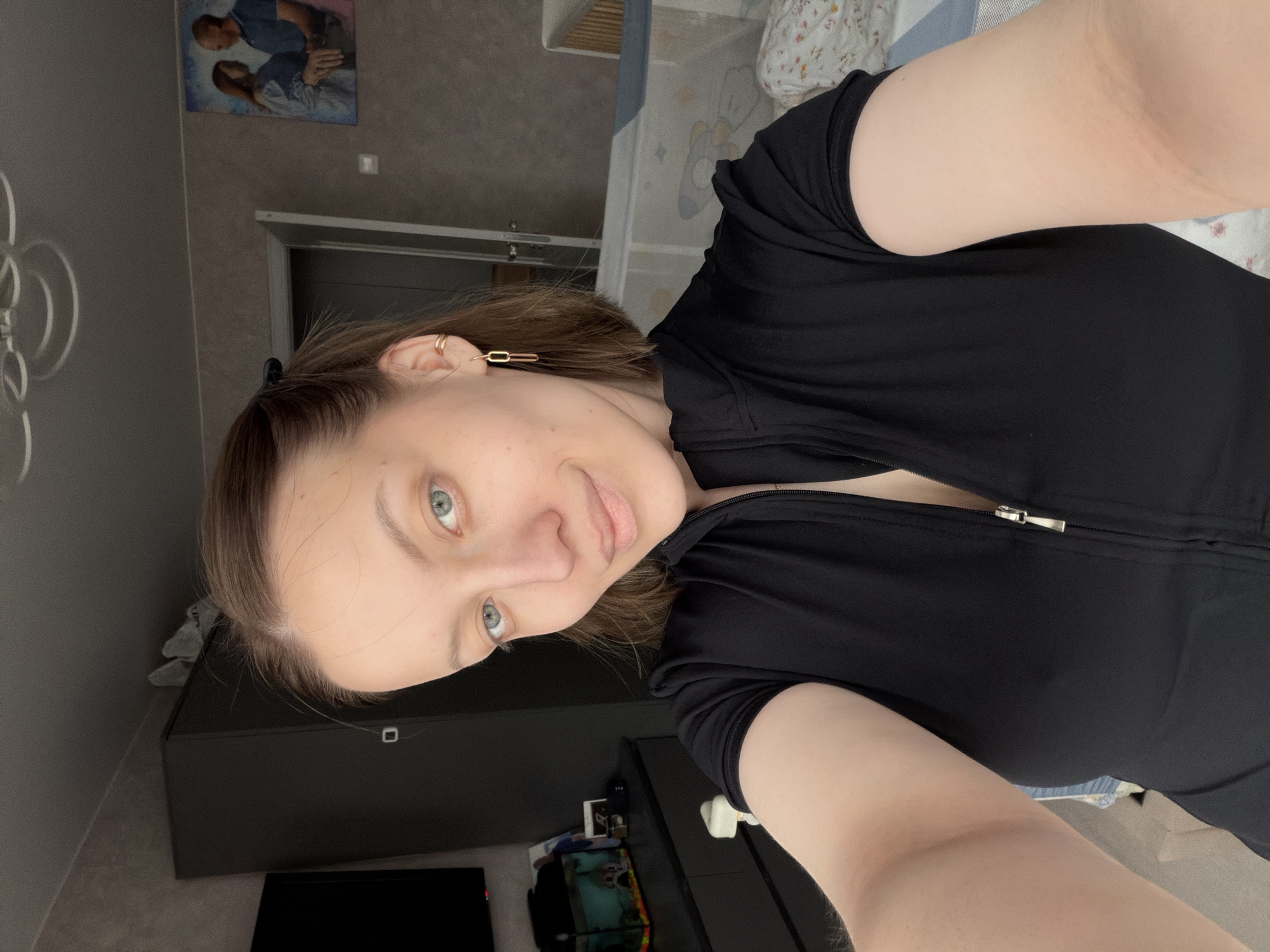 An indoor selfie captures a woman looking directly at the camera with a slight smile. She has light-colored eyes (blue/green) and brown hair. Her left ear is adorned with two small hoop piercings on the upper cartilage and a gold-toned, chain-link style earring dangling from the lobe. She is wearing a black, short-sleeved top with a visible zipper closure.
The setting appears to be a room, possibly a bedroom or living space in Reshetnikovo, Russia. In the background, to the woman's left, is a dark dresser or cabinet with a television screen below it. Above this, on a gray wall, hangs a framed picture depicting two individuals, a man and a woman, in what appears to be an embrace. To the woman's right, part of a bed with light-colored, patterned bedding is visible. The lighting suggests it is daytime. There is no visible text in the image.