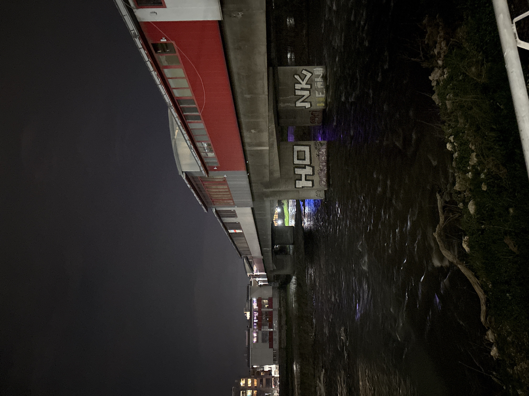 The image, taken at night in Plovdiv, Bulgaria, shows the Maritza River reflecting city lights. Graffiti is visible on a concrete bridge support with the letters "HO" and "NK". A building with red accents and multiple windows runs parallel to the river's edge. On the opposite side of the river, vegetation grows along the bank. In the distance, buildings with illuminated windows rise into the night sky. The water surface is turbulent, with bright streaks caused by light reflecting off of it. A white pole stands at the right edge of the picture.