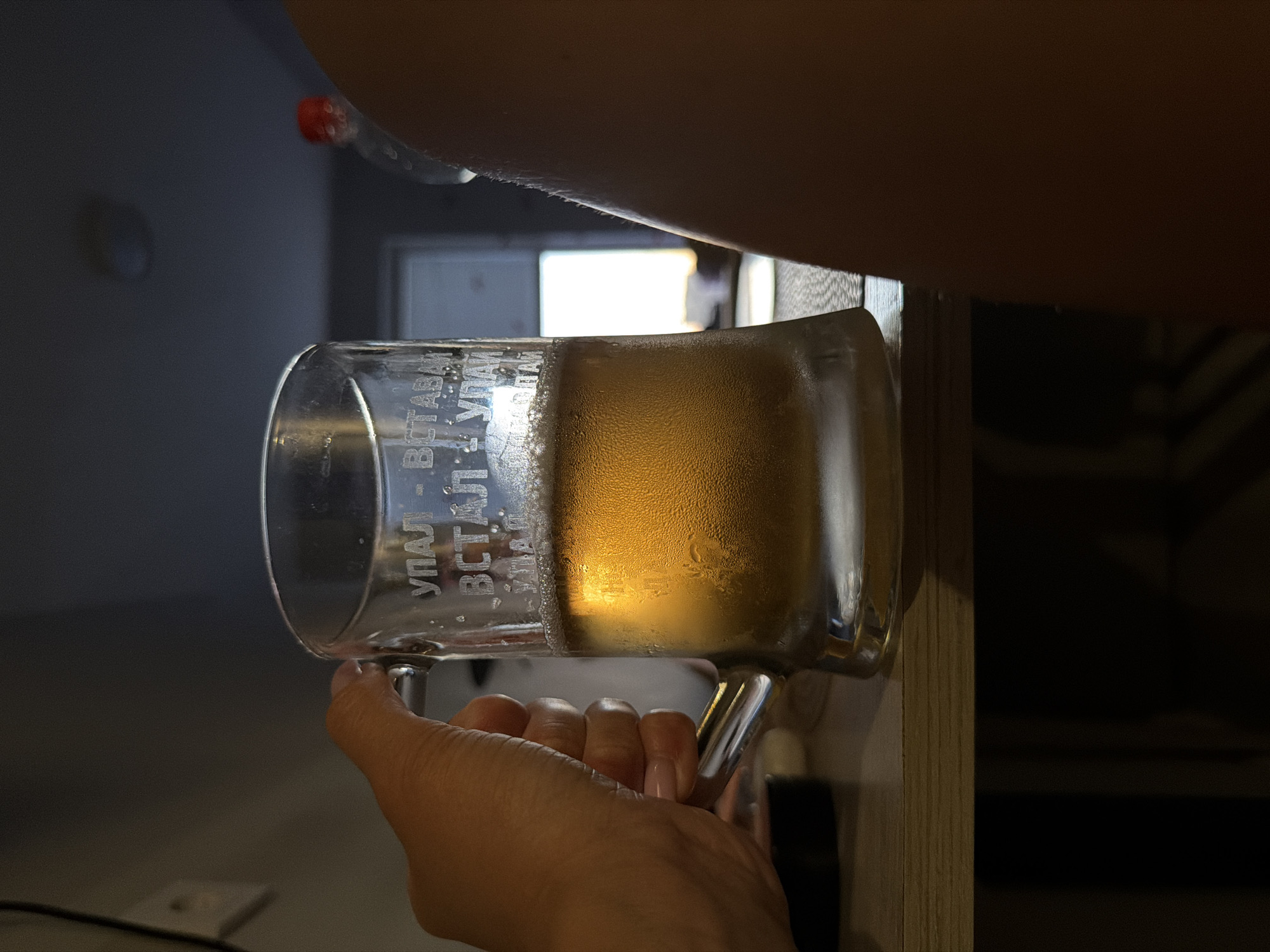The image shows a hand holding a glass mug filled with a golden liquid, likely beer. The mug has frosted glass and appears to be engraved with text, though the words are difficult to decipher clearly. Some of the visible letters appear to be Cyrillic. The liquid inside the mug has condensation on the outside of the glass, suggesting it is cold. The background is blurred, but hints at an indoor setting with soft lighting and what might be a window or light source in the distance. The image is taken from a low angle, looking up at the mug and hand. The lighting suggests it could be daytime, given the brightness in the background. The overall mood is casual and relaxed, perhaps depicting someone about to enjoy a drink.