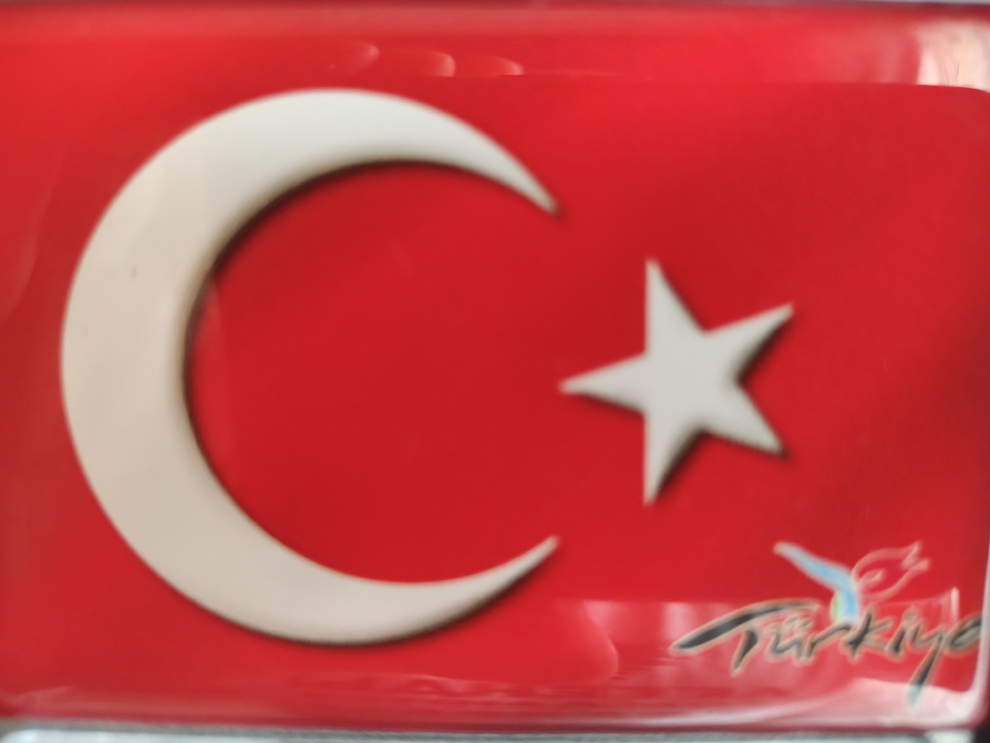 The image shows a close-up, slightly blurred view of a red rectangular object with the Turkish flag emblem. The emblem consists of a white crescent moon and a white star on the red background. In the lower right corner, there is a stylized inscription that appears to be "Türkiye". The object seems to be reflective, as suggested by the highlights on the surface. The location in the city of Придорожный, Russia is not discernible from the provided image.