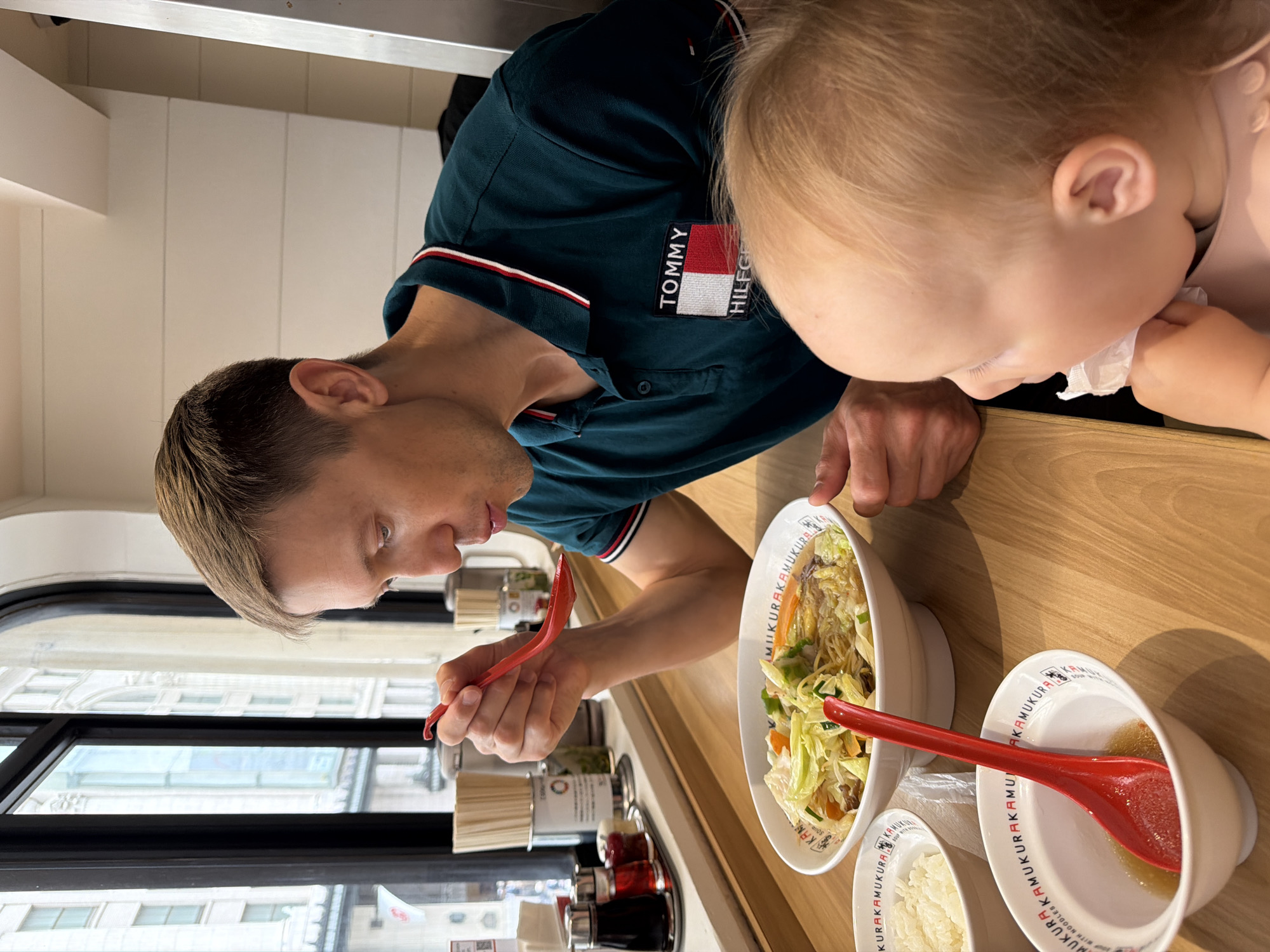 An adult male and a young child are seated at a wooden counter in a Kamukura Ramen restaurant, situated in an urban area consistent with the city of Osaka, Japan. The male, with light-colored short hair and wearing a dark teal polo shirt featuring a Tommy Hilfiger logo, is captured mid-action, bringing a red plastic ramen spoon filled with food to his mouth from a large white bowl of ramen. The ramen bowl contains noodles, broth, chopped cabbage, carrots, and other vegetables. To his left, a smaller white bowl holds plain white rice, and another partially empty small white bowl contains some liquid and a resting red plastic ramen spoon. A blonde-haired toddler, wearing a pink bib, is partially visible on the right side of the frame, holding a crumpled white napkin near their mouth. In the background, behind the counter, a large arched window reveals a view of multi-story city buildings and a visible Japanese flag on a pole. Various restaurant condiments and chopstick holders are arranged on the counter behind the individuals.