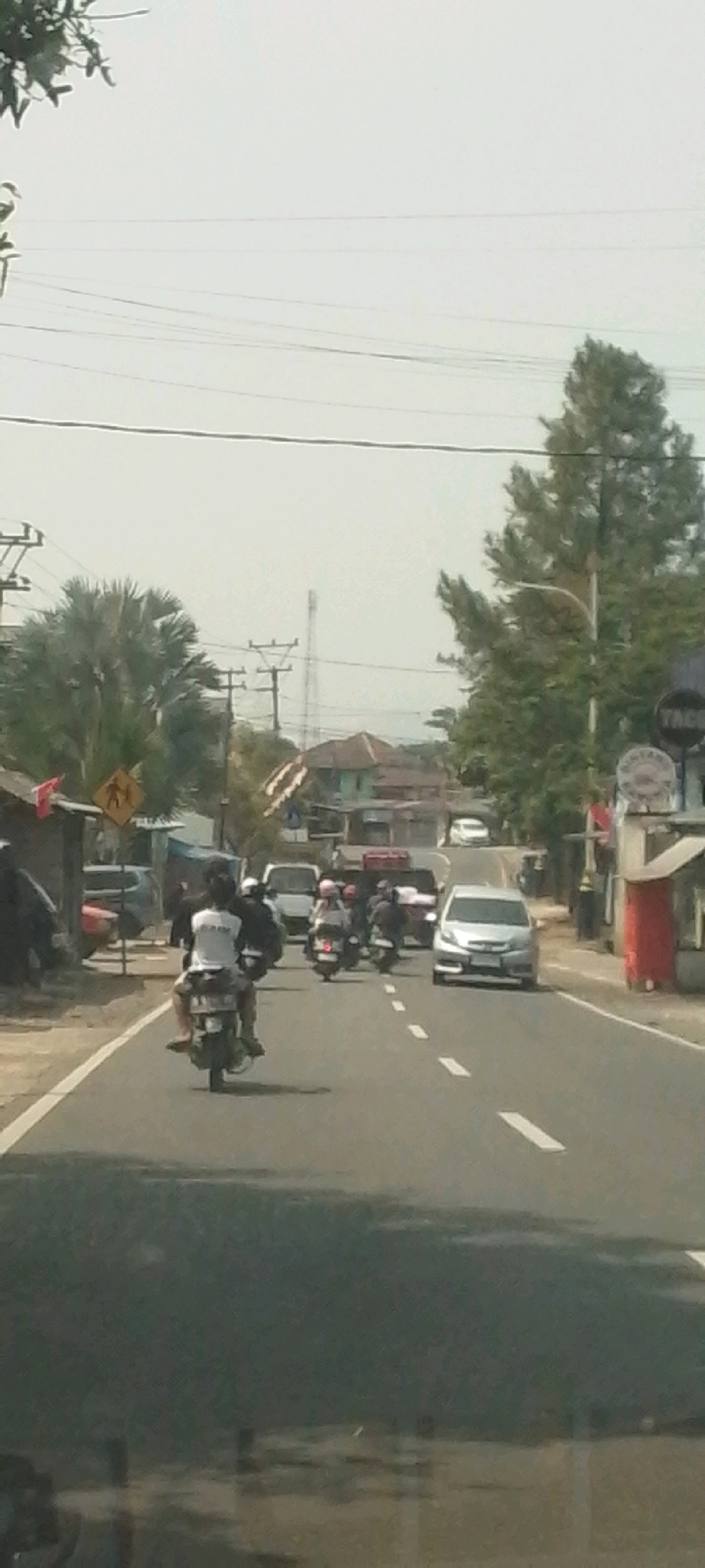 The image depicts a road in the city of Peuteuycondong, Indonesia, on a bright, overcast day. The road is paved and marked with white dashed lines. Several vehicles are present, including motorcycles and cars. On the left side of the road, there is a pedestrian crossing sign with a yellow diamond shape and a silhouette of two people walking. Buildings line the side of the road, and there are trees and utility poles visible in the background. A few individuals are seen riding motorcycles, some with passengers. The overall scene suggests a typical day on a moderately busy road in a residential or semi-urban area.