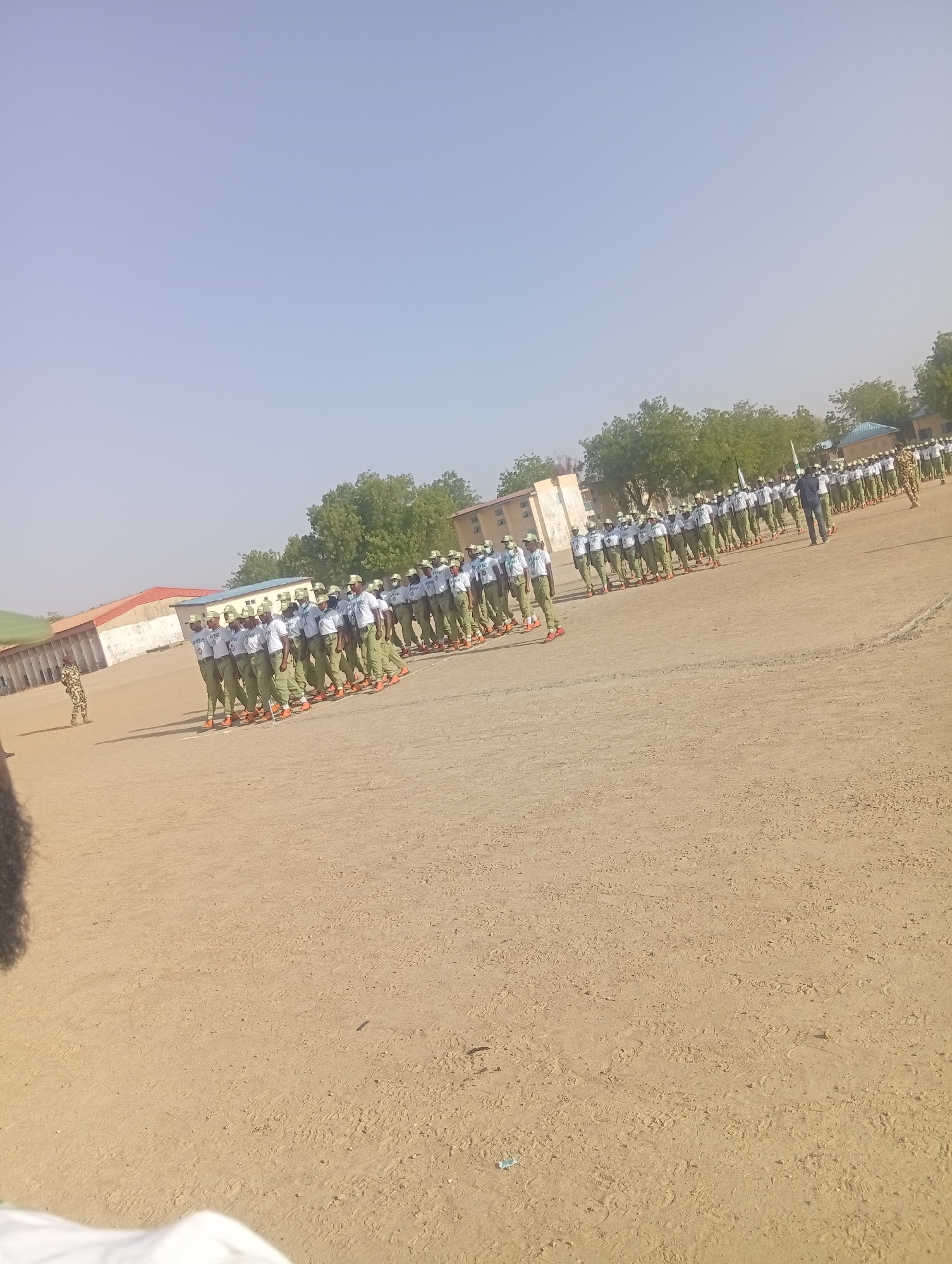 A large group of people in matching white shirts, green pants, and orange shoes are standing in rows on a dirt field. There are buildings and trees in the background, under a clear, blue sky. The location appears to be Maiduguri, Nigeria.