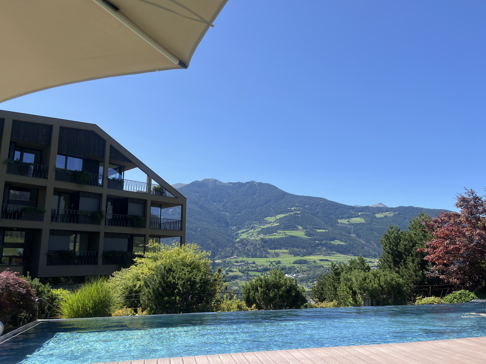 An eye-level, wide shot captures a serene outdoor scene with a pool in the foreground and a mountain landscape in the background. The pool, filled with clear, shimmering blue water, is framed by a wooden deck. Beyond the pool, lush green trees and bushes create a verdant border. In the mid-ground, a modern, multi-story building with dark brown siding and balconies stands against the backdrop of rolling green hills dotted with small structures. The furthest background is dominated by a majestic, tree-covered mountain range under a clear, bright blue sky. The scene is bathed in bright sunlight, suggesting a warm, pleasant day. There are no people visible in the frame. The location is the city of Brixen - Bressanone, Italy.