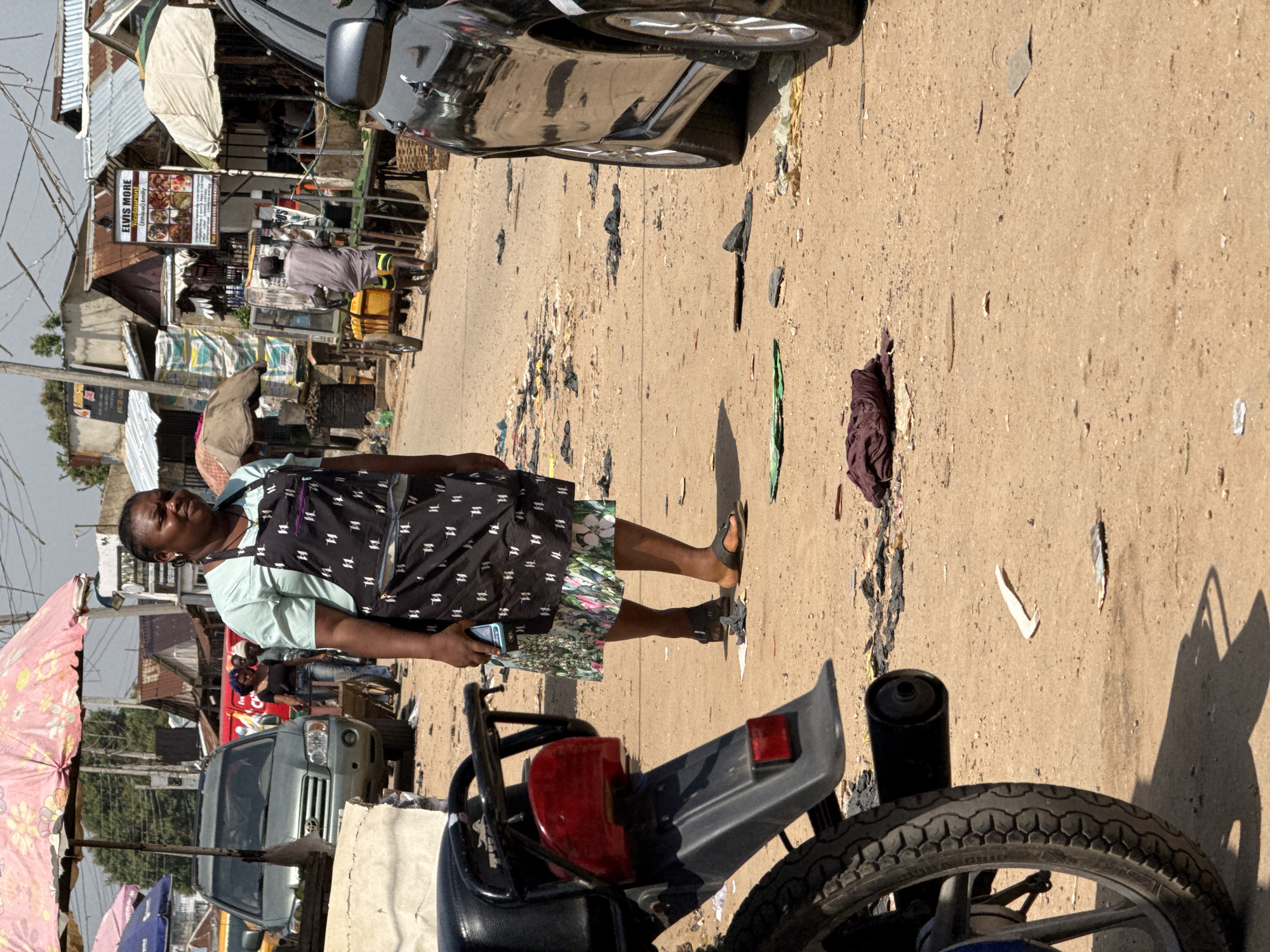 The image depicts a street scene in Abuja, Nigeria, characterized by a dusty, unpaved road lined with various structures and vehicles. In the foreground, a portion of a black motorcycle is visible on the left, with its rear tire and exhaust pipe in sharp focus. A woman, appearing to be of African descent, stands in the middle ground, facing slightly to her right. She is wearing a light blue top and a black apron with a white pattern. She is holding a mobile phone in her right hand. Behind her, a grey SUV is parked on the left side of the road. Further back, several market stalls or informal shops with corrugated metal roofs are visible. A man in a grey garment is pushing a cart laden with yellow containers down the road. A black car is parked on the right side of the image, obscuring part of the background. The ground is sandy and scattered with debris, including plastic bags and fabric. The scene suggests a bustling outdoor market or a roadside commercial area.