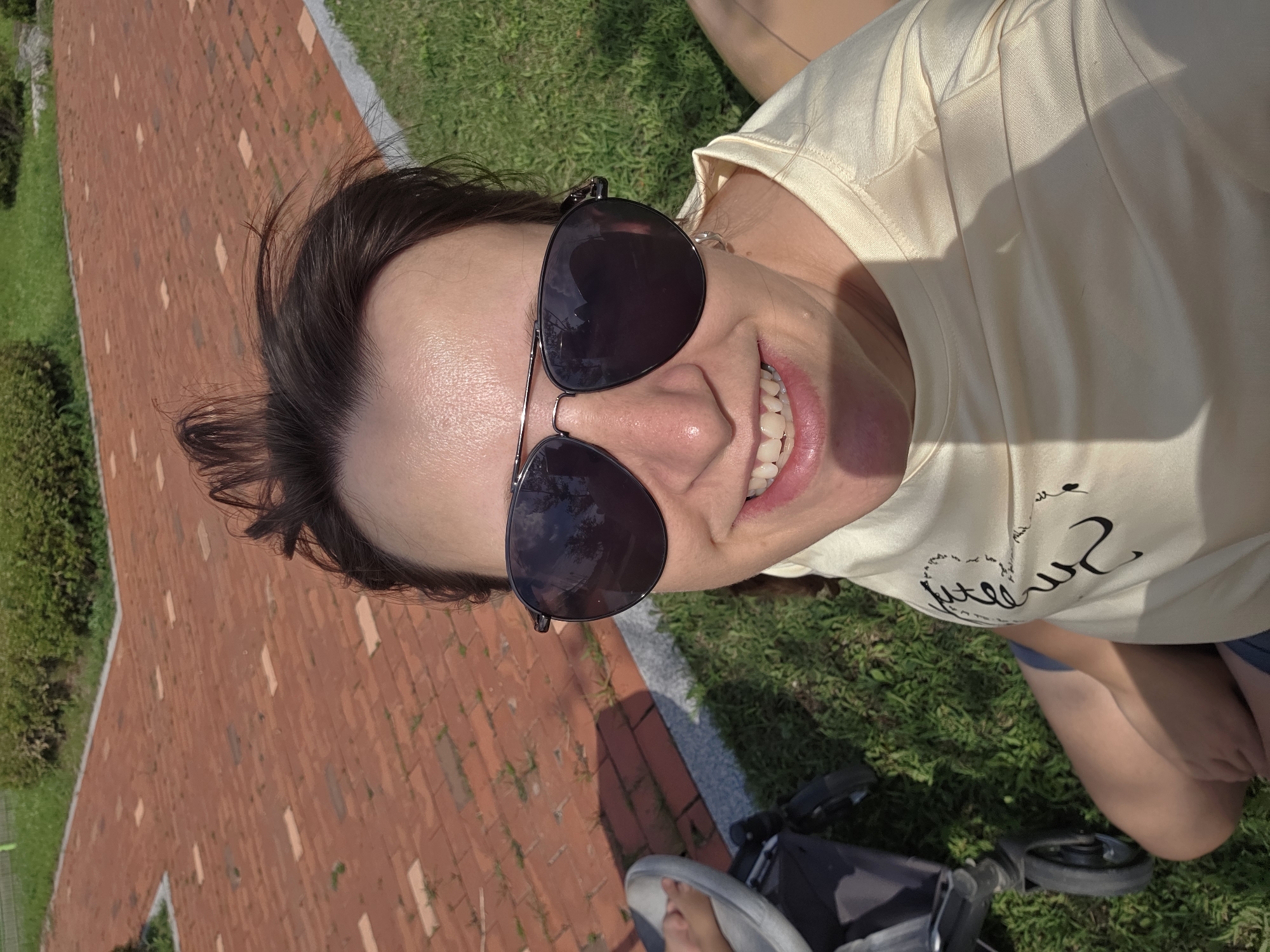 A person wearing sunglasses and a light-colored t-shirt is smiling at the camera outdoors. The person's hair is dark brown and pulled back. The background shows a paved area with red bricks, green grass, and some bushes. A stroller is visible to the left of the person. The scene appears to be in a park or public outdoor space. The timestamp indicates the image was taken in Dunnae-myeon, South Korea.