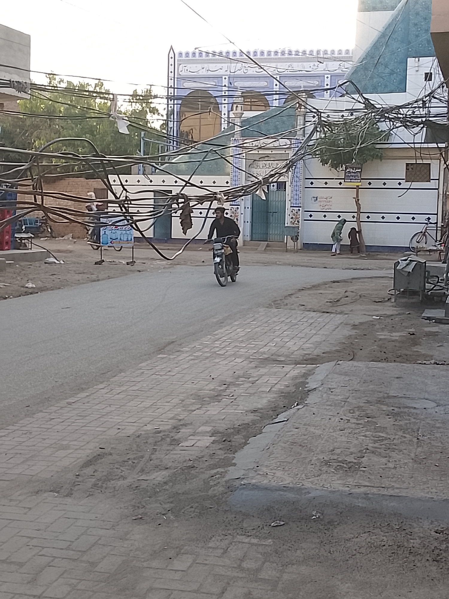 The image depicts a daytime street scene, likely in a town or village within a South Asian or Middle Eastern setting. In the foreground, a man is riding a dark motorcycle down a paved road, wearing dark clothing and a light-colored cap or turban. The road consists of both asphalt and a brick-paved section on the right, with dusty, uneven ground at the edges.
Above the street, a dense, complex network of black electrical wires stretches across the frame, obscuring parts of the background. In the mid-ground, a prominent building with a light blue and white facade dominates the scene. This structure, possibly a mosque or religious institution, features intricate calligraphic script (likely Urdu or Arabic) and geometric patterns above arched entrances.
Several other people are visible in the background. To the left of the main building, a person stands near a sign advertising "Clean Water" (صاف پانی) in Urdu, with a graphic of a water bottle. On the right, two figures are near a building with a white wall adorned with black diamond patterns; one appears to be a woman in a green outfit, and the other is a smaller individual. A bicycle is parked against this patterned wall, and a metal utility box is also present on the right. On the far left, red crates are stacked.
The sky is bright and clear, indicating fair weather during daylight hours, likely late morning or afternoon given the soft lighting. The dusty ground, distinctive architecture, and extensive overhead wiring are notable location cues.