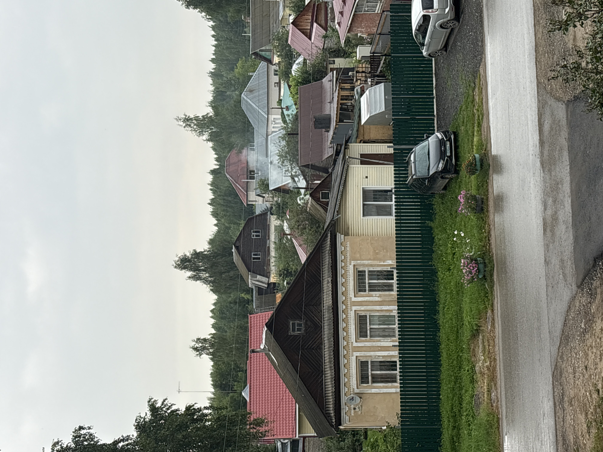 The image depicts a street view in Krasnokamsk, Russia, on an overcast day. In the foreground, a wet asphalt road reflects the cloudy sky. To the right of the road is a gravel driveway with two cars parked: a silver hatchback and a dark-colored SUV. A green wooden fence runs along the edge of the property, separating it from a grassy area. Behind the fence, a row of houses with various roof colors and architectural styles is visible. Some houses have chimneys, and a faint plume of smoke can be seen rising from one of them. The houses are surrounded by trees and vegetation. The overall scene suggests a residential area in a rural or suburban setting.
