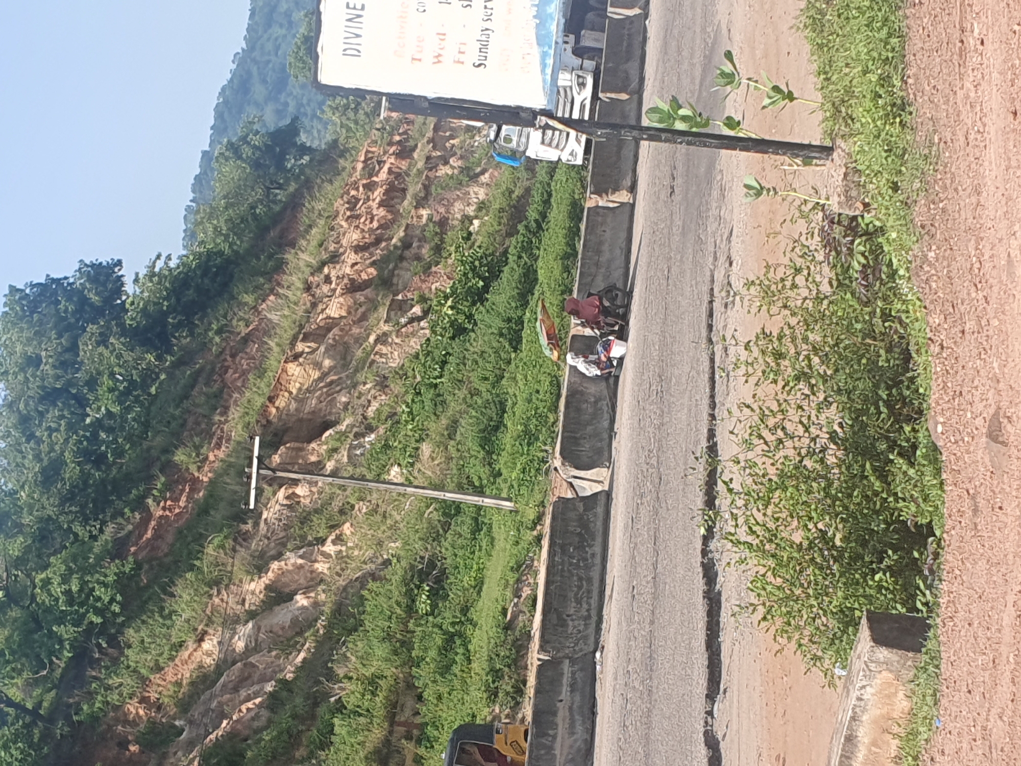 This image depicts a roadside scene in Lokoja, Nigeria. A person is sitting on a bicycle next to a pile of belongings, including what appears to be a colorful umbrella or tarp and a white bag. The person is wearing a dark red top. The scene is set alongside a paved road with a concrete barrier. Beyond the road, there is a steep, rocky hillside covered in lush green vegetation. A utility pole with wires runs parallel to the road. In the background, a sign with the word "DIVINE" is visible, along with some text that appears to list days of the week and services. The sky is clear and blue, suggesting a bright, sunny day. The overall impression is of a rural or semi-urban roadside environment.