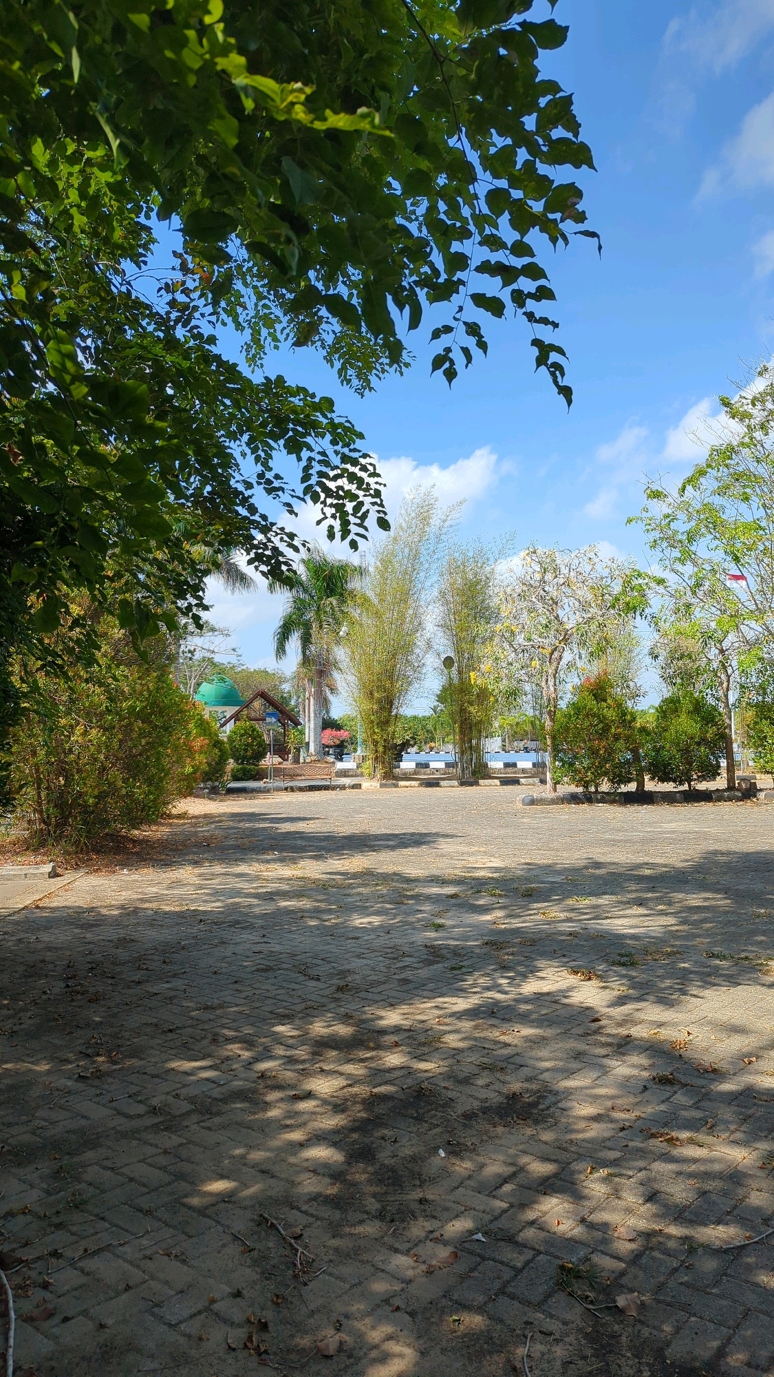 In Gunungtinggi, Indonesia, an outdoor paved area is shown under a bright, partially cloudy sky. The paving stones are laid in a herringbone pattern and are partially covered with fallen leaves and twigs. Shadows from trees are cast across the ground, creating a dappled effect. In the background, several vehicles are parked, and a variety of trees and vegetation are visible. To the left, a green-domed building, possibly a mosque, is partially obscured by trees. Tall bamboo stalks and palm trees add to the tropical atmosphere. The scene appears to be a public space, perhaps a parking lot or courtyard adjacent to a facility.