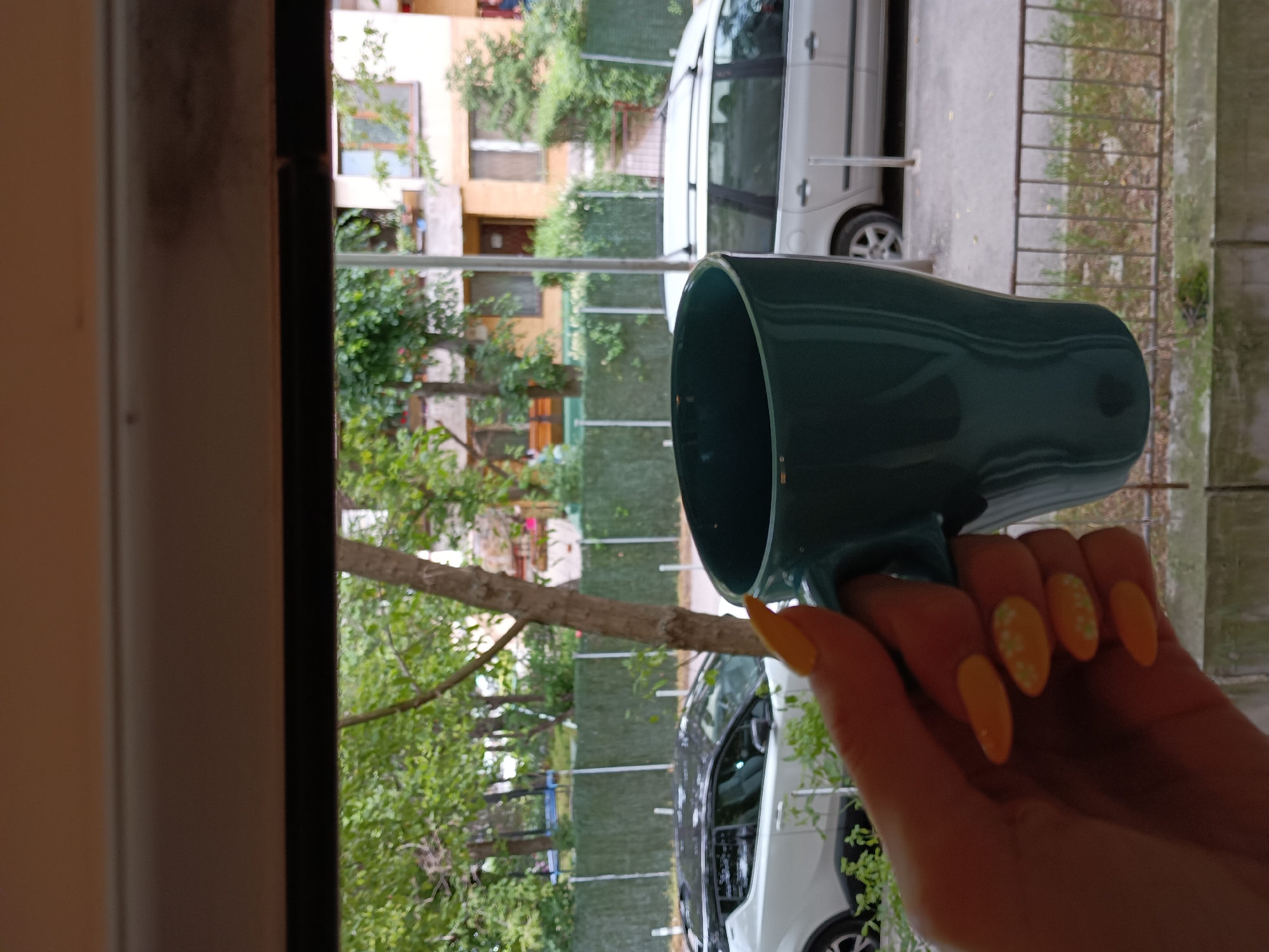 The image presents a street-level view from an elevated position, likely a window or balcony, within a residential area of Sofia, Bulgaria. In the immediate foreground, a human hand, featuring almond-shaped nails painted bright yellow-orange with white floral decals on the ring and middle fingers, grips a glossy dark teal ceramic mug. The outdoor scene reveals a paved street bordered by a prominent green slatted fence, supported by white posts. Beyond the fence, a dense canopy of green trees obscures parts of several multi-story residential buildings, which are ochre and beige in color with visible balconies and windows. Two vehicles are parked along the street: a white modern two-door car with a black roof on the left, and a silver station wagon equipped with roof rails on the right. A dark metal railing surmounts a concrete ledge in the bottom foreground, exhibiting some moss and debris. The scene is illuminated by diffused natural light, suggesting an overcast day.