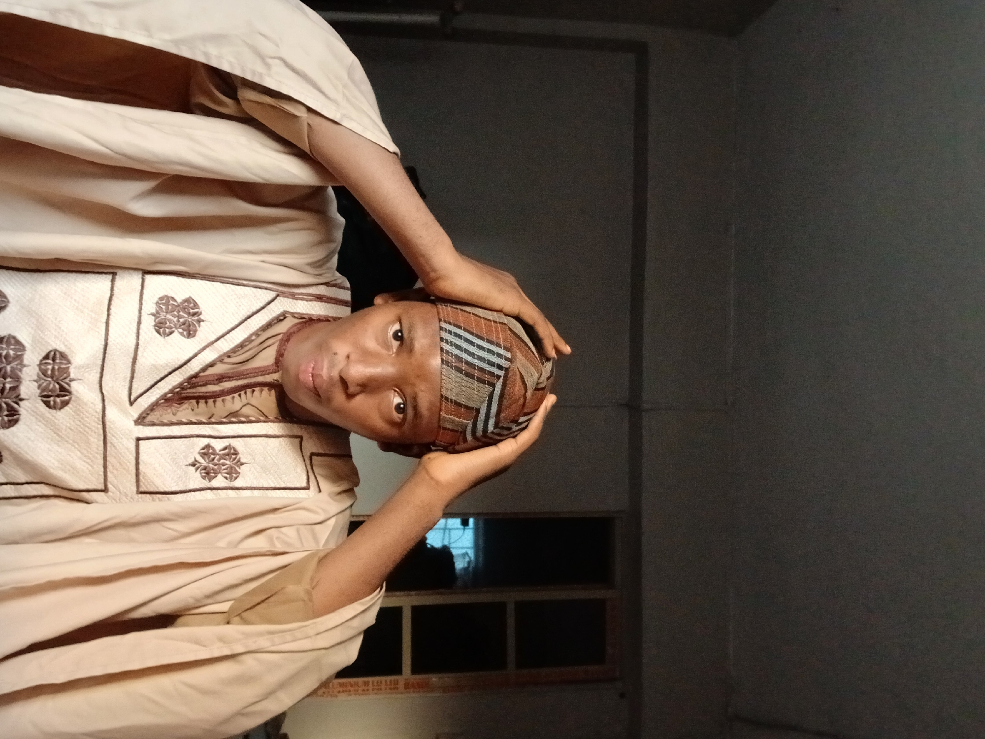 A person wearing a traditional brown and white patterned garment and a matching cap is visible in a dimly lit indoor space. The person is positioned so that their face is visible, and their hands are touching the cap on their head. Behind the person, there is a window in the distance and a plain wall. The photograph appears to be taken inside what might be a home or office. The information available does not allow for determination of the exact location within Lagos, Nigeria.