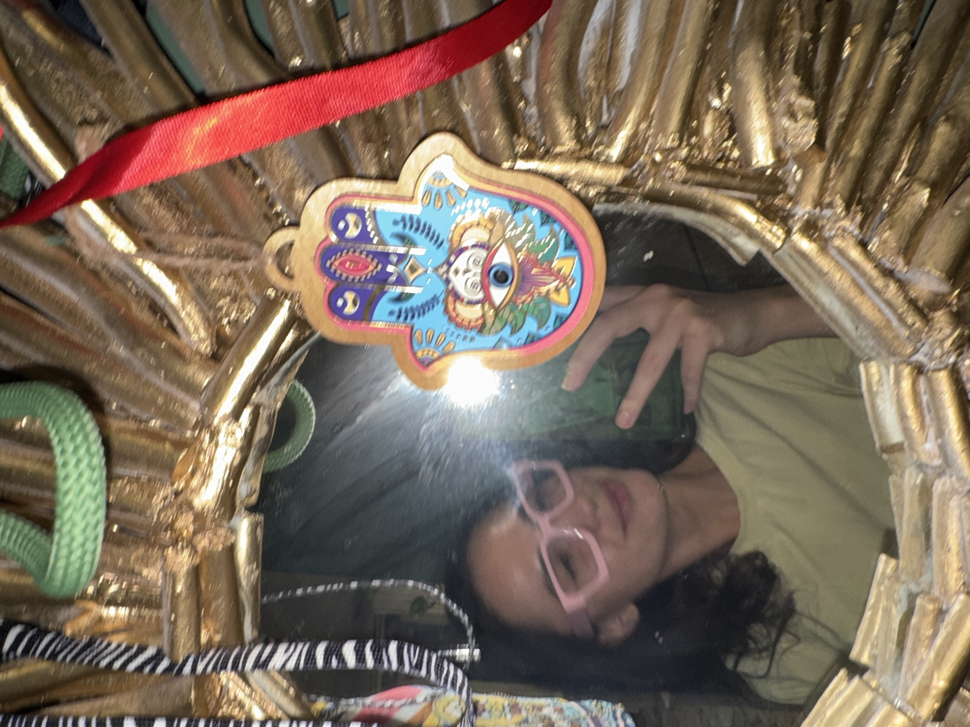A person with pink glasses is reflected in a mirror framed in gold. An amulet in the shape of a hand with a design of blue, white, and gold is hanging at the top of the mirror frame. A red ribbon is above the hand. A green rope and a zebra-striped pattern are also partially visible on the frame. The location is not discernible from the image.