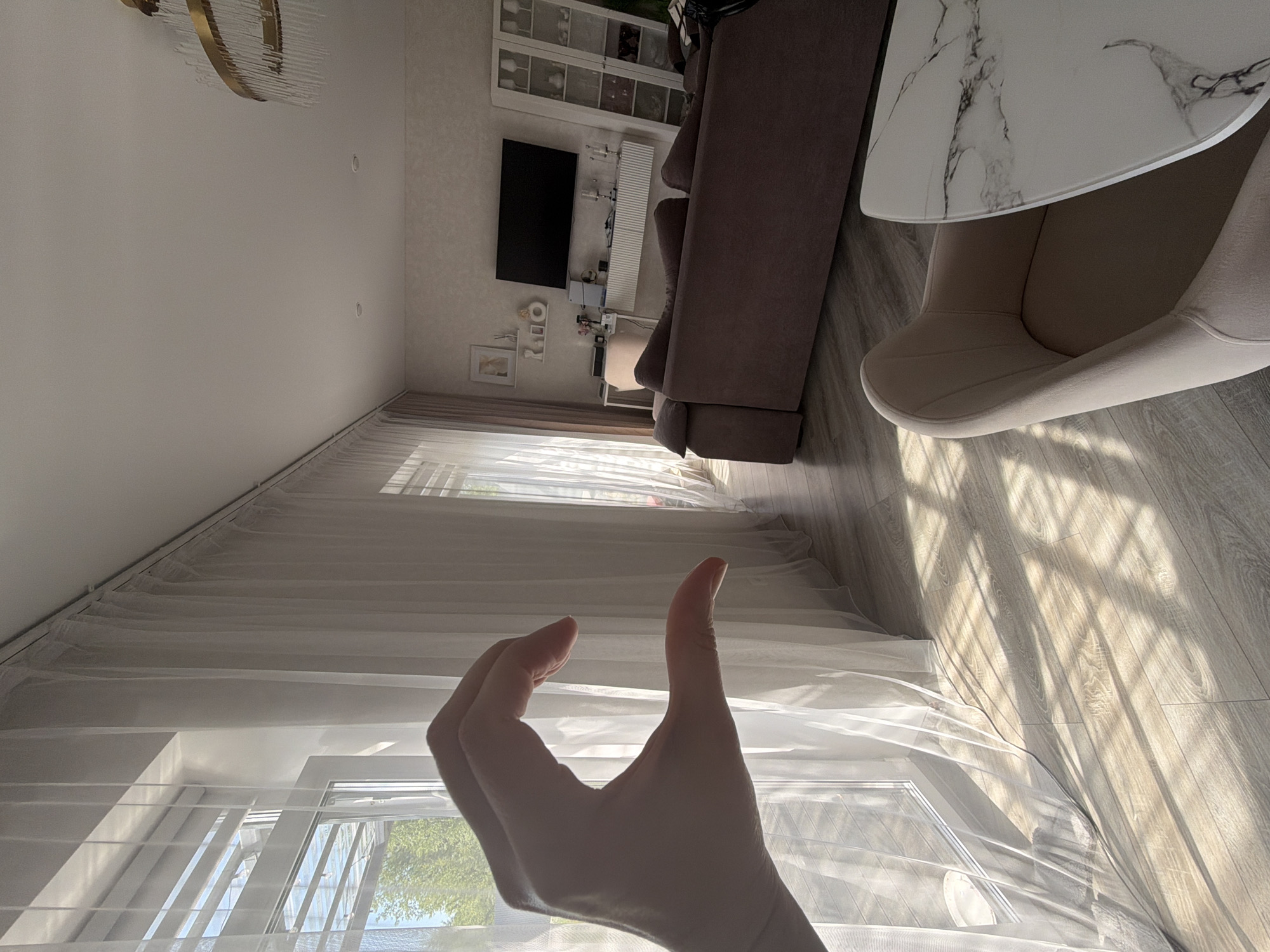 The image depicts a sunlit interior of a living room, likely in Malinovka, Russia. The foreground features a hand forming a heart shape, partially obscuring a window with sheer white curtains. Sunlight streams through the window and curtains, casting geometric shadows on the light-colored wooden floor.
The background reveals a modern living space with a large, dark-colored sectional sofa. Adjacent to the sofa is a sleek white entertainment console mounted on the wall, supporting a flat-screen television. Above the console, a minimalist shelf displays decorative items, including framed pictures and a vase. To the right, a tall white glass-fronted cabinet, possibly a display cabinet, stands against the wall, containing various glassware and decorative objects. A single, light-colored upholstered chair is visible in the lower right corner, next to a marble-topped table. A contemporary chandelier with a golden frame and glass elements hangs from the ceiling. The overall impression is one of a well-decorated and tidy residential interior.