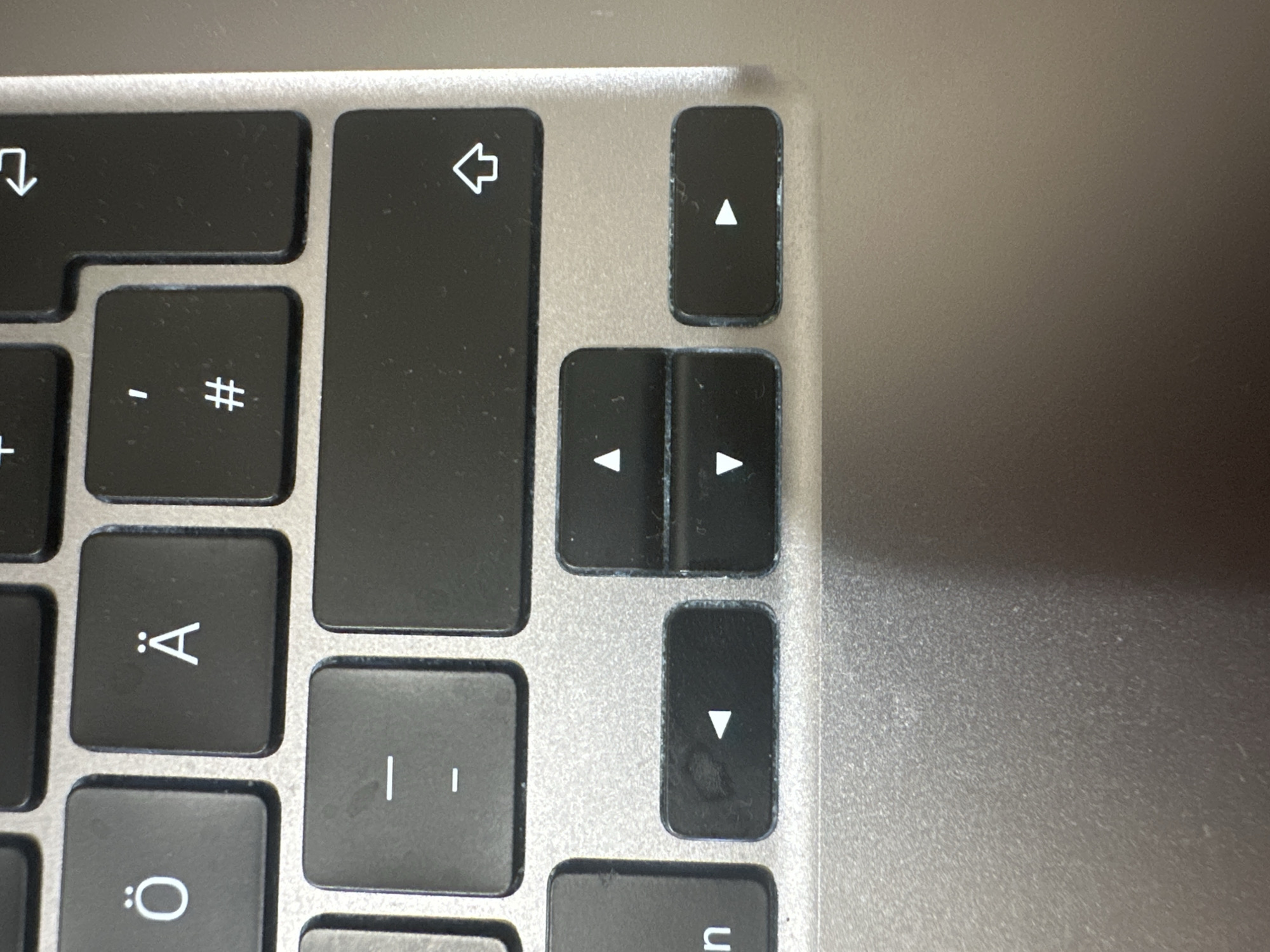 The image is a close-up, high-angle shot of a section of a laptop keyboard, likely a MacBook based on the key design and layout. The keyboard is silver and appears to be made of aluminum. Several keys are visible, including special character keys like 'Ö' and 'Ä', a dash key, a key with a '#' symbol, and arrow keys (left, up, down, and right). The keys are black with white symbols. Some keys, particularly the arrow keys and the key to the left of the arrow keys, show signs of wear and smudging, indicating use. The lighting suggests an indoor environment, possibly a desk or table. There are no people visible in the image. The background is out of focus and shows the textured surface of the laptop. The image does not contain any information about the city of Hamburg, Germany.