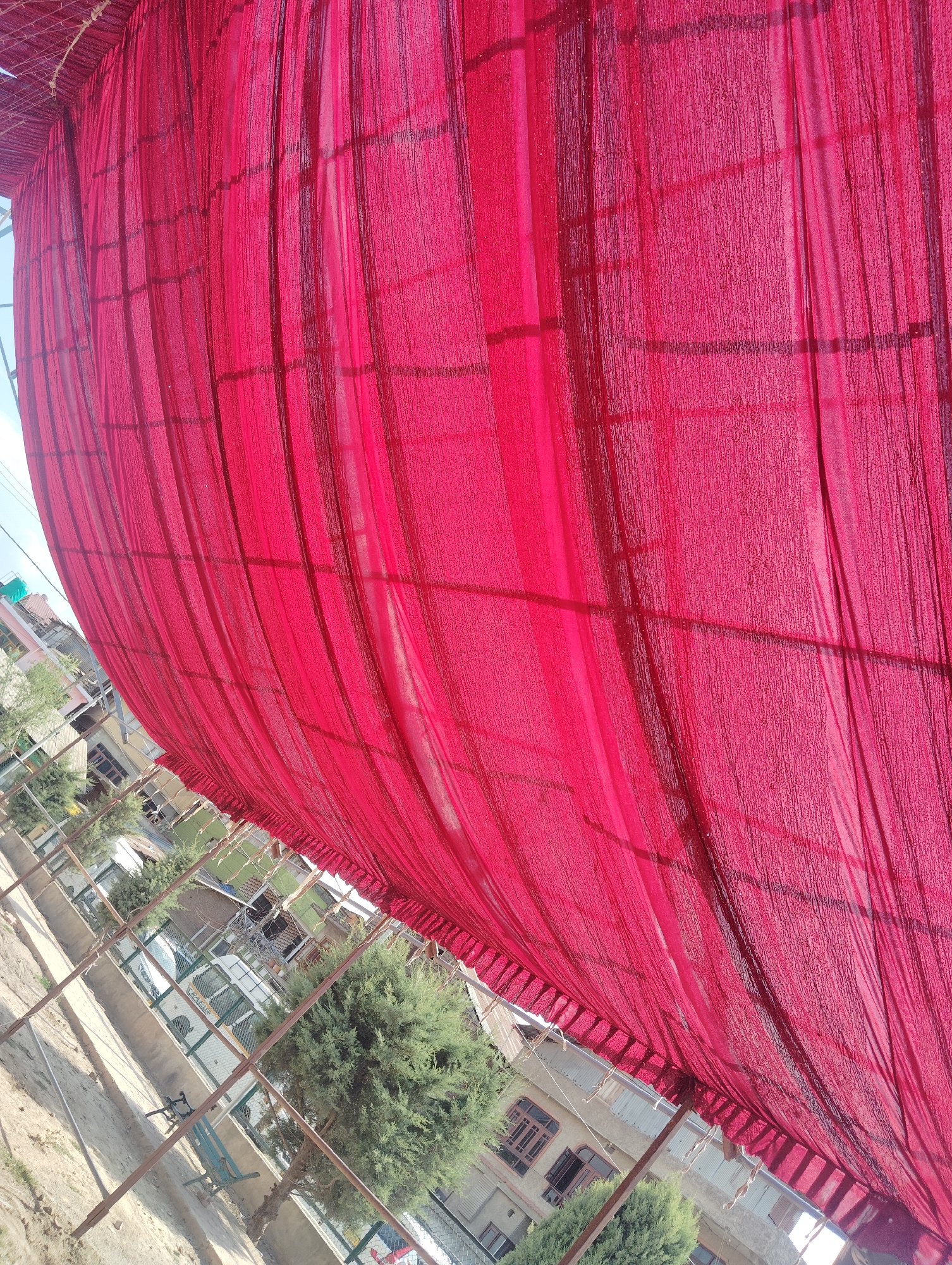 A low-angle shot of a large, draped red fabric is presented, angled upwards towards a bright sky. The fabric appears to be part of a temporary structure, possibly a tent or canopy, supported by metal poles that are visible in the foreground and midground. The texture of the fabric is visible, with vertical pleats and a grid-like pattern of darker threads.
Beneath the fabric, a residential area is visible. Buildings with multiple windows and balconies are apparent, along with trees and a white vehicle. The architecture suggests an urban or semi-urban setting. The overall impression is that of an outdoor event or construction site, likely in the city of Srinagar, India, given the architectural style and vegetation. The scene is illuminated by bright sunlight, casting shadows on the ground and highlighting the texture of the red fabric. No people are visible in the frame.
