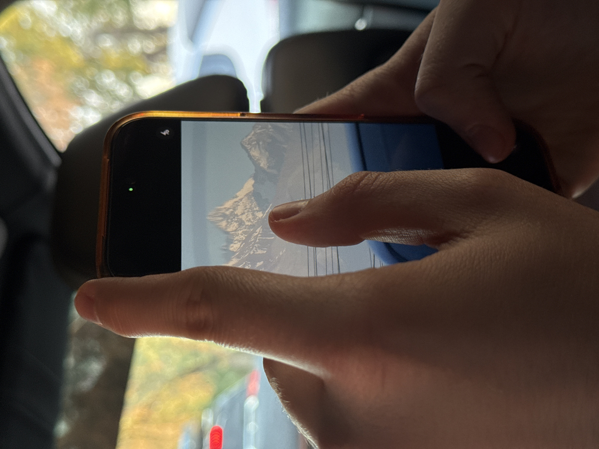 A person is holding a smartphone horizontally, with their thumbs and fingers visible. The smartphone screen displays an image of a mountain range. The background of the scene is blurred, suggesting it is either a car interior or a room with a window. Outside the window, the scenery is out of focus, showing greenery and some bokeh lights, indicating it might be daytime. The phone's camera is likely in use, as a small green dot is visible on the screen, which often signifies camera or microphone usage. The overall impression is that the person is capturing or viewing a photograph of a scenic mountain landscape.