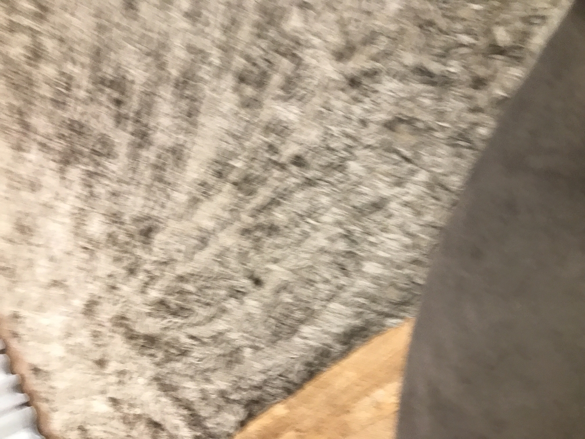 The image shows a close-up of a textured surface, possibly a carpet or rug, with a mottled gray and beige color pattern. A portion of a smooth, dark gray object is visible on the right side of the image, partially obscuring the textured surface. A segment of a light-brown, possibly wooden, object is visible in the lower center of the frame. The image is blurry and lacks sharp details. There is no indication of a location in Yerevan, Armenia or any human presence or actions.