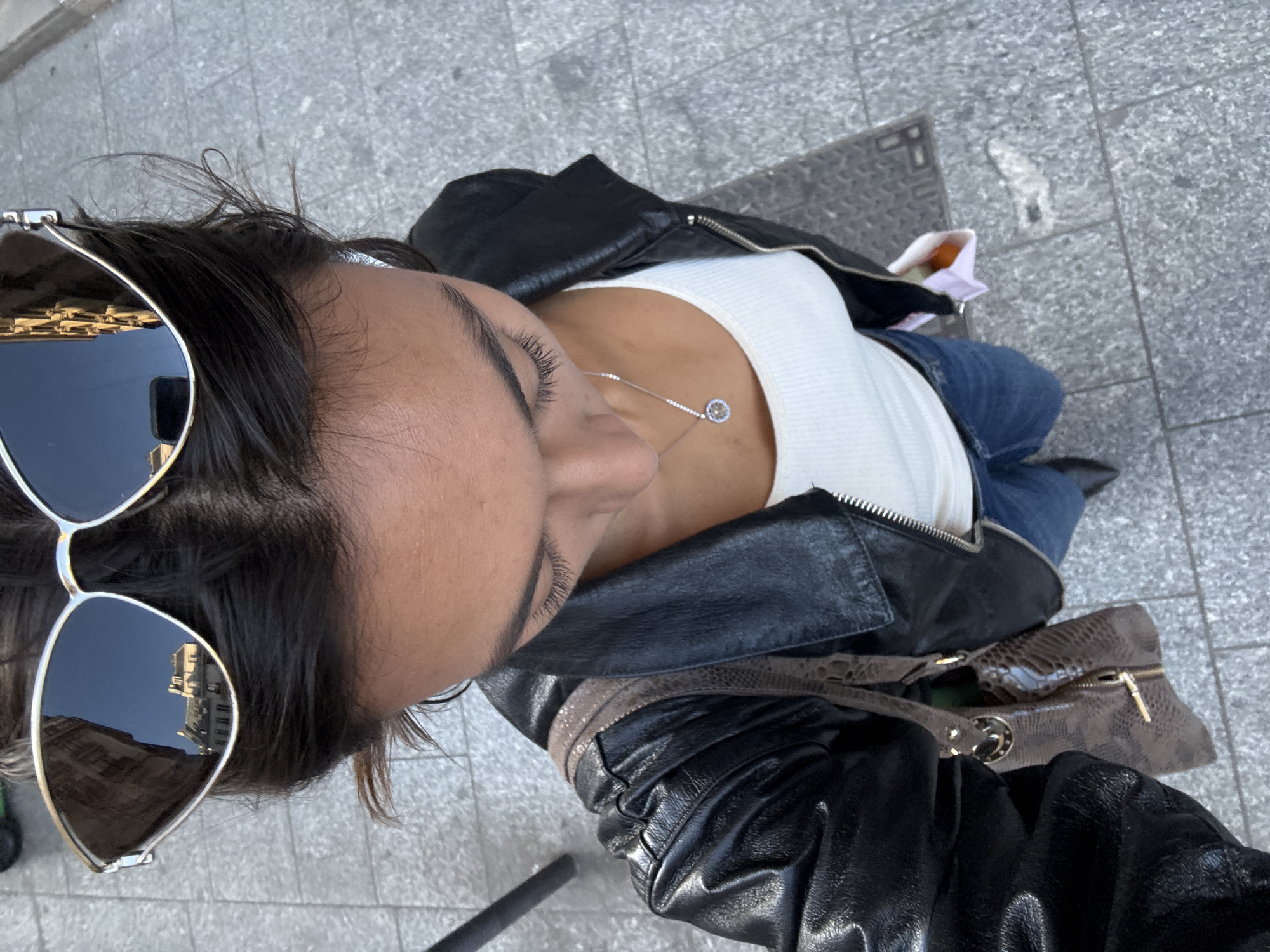 The image presents a high-angle, downward-looking view of a person outdoors. The subject has dark hair, which is slightly disheveled on top, and dark, well-defined eyebrows. Large, reflective sunglasses with light-colored frames are perched on their head, obscuring the upper part of their face. The reflections in the sunglass lenses show a building with multiple windows under a clear blue sky, indicative of daytime.
The person is wearing a black leather jacket with visible zippers on the shoulders, layered over a white ribbed tank top or camisole. A delicate silver chain necklace with a circular pendant, featuring a central stone surrounded by smaller ones, is visible on their chest. Parts of blue jeans are seen below the jacket. A brown handbag with a textured, possibly faux crocodile or snakeskin, pattern and gold hardware (including a zipper and a decorative ring) is positioned to the person's right. Additionally, a small white bag containing an orange-brown item is seen near their left hip.
The ground beneath the person is paved with grey, rectangular tiles, common in urban settings like sidewalks or public squares. A metal utility grate is also visible on the ground. The bright light and clear sky reflected in the sunglasses suggest sunny weather. The architectural style reflected in the lenses, coupled with the contextual information, points to a European city, consistent with Paris, France. There is no visible text in the image.
